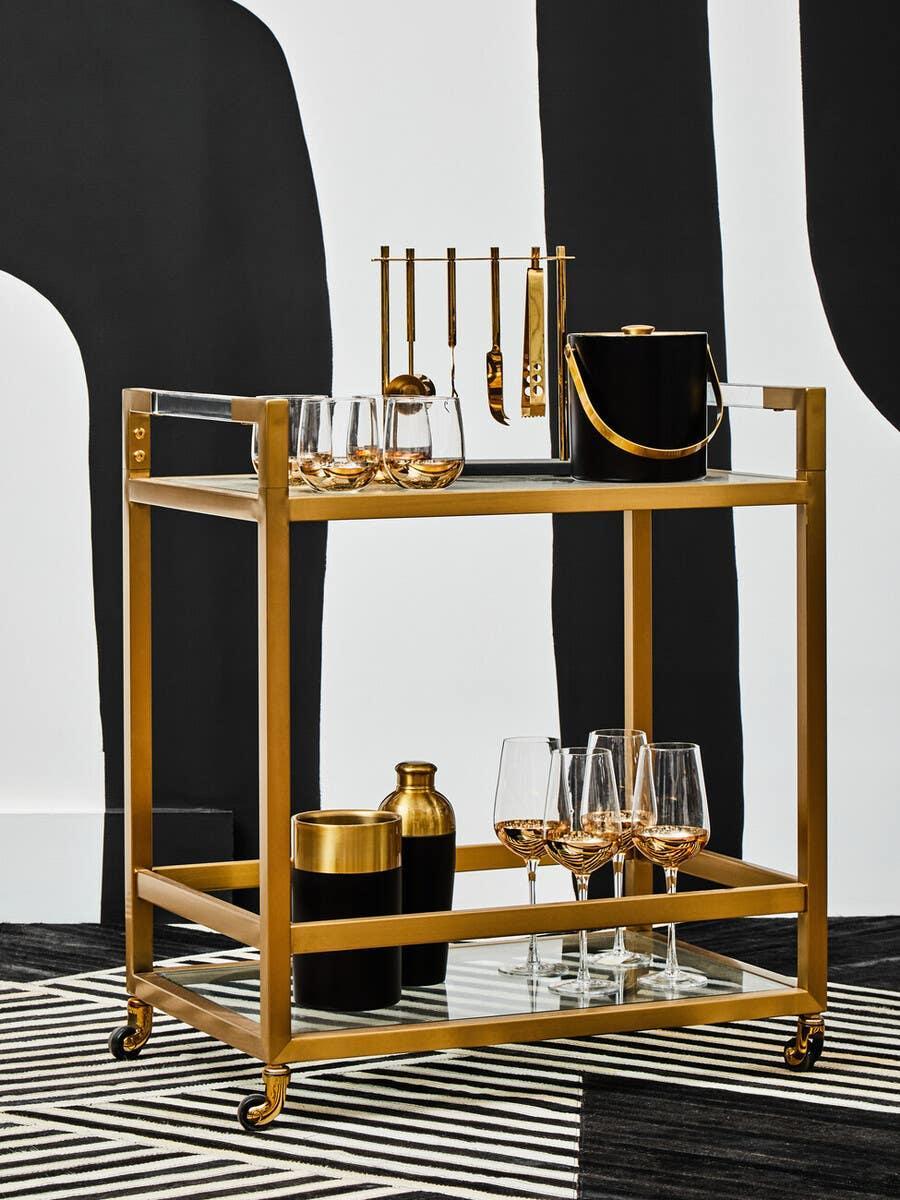 Gold metal bar cart with black tray, glassware, cocktail tools, and ice bucket on wheels