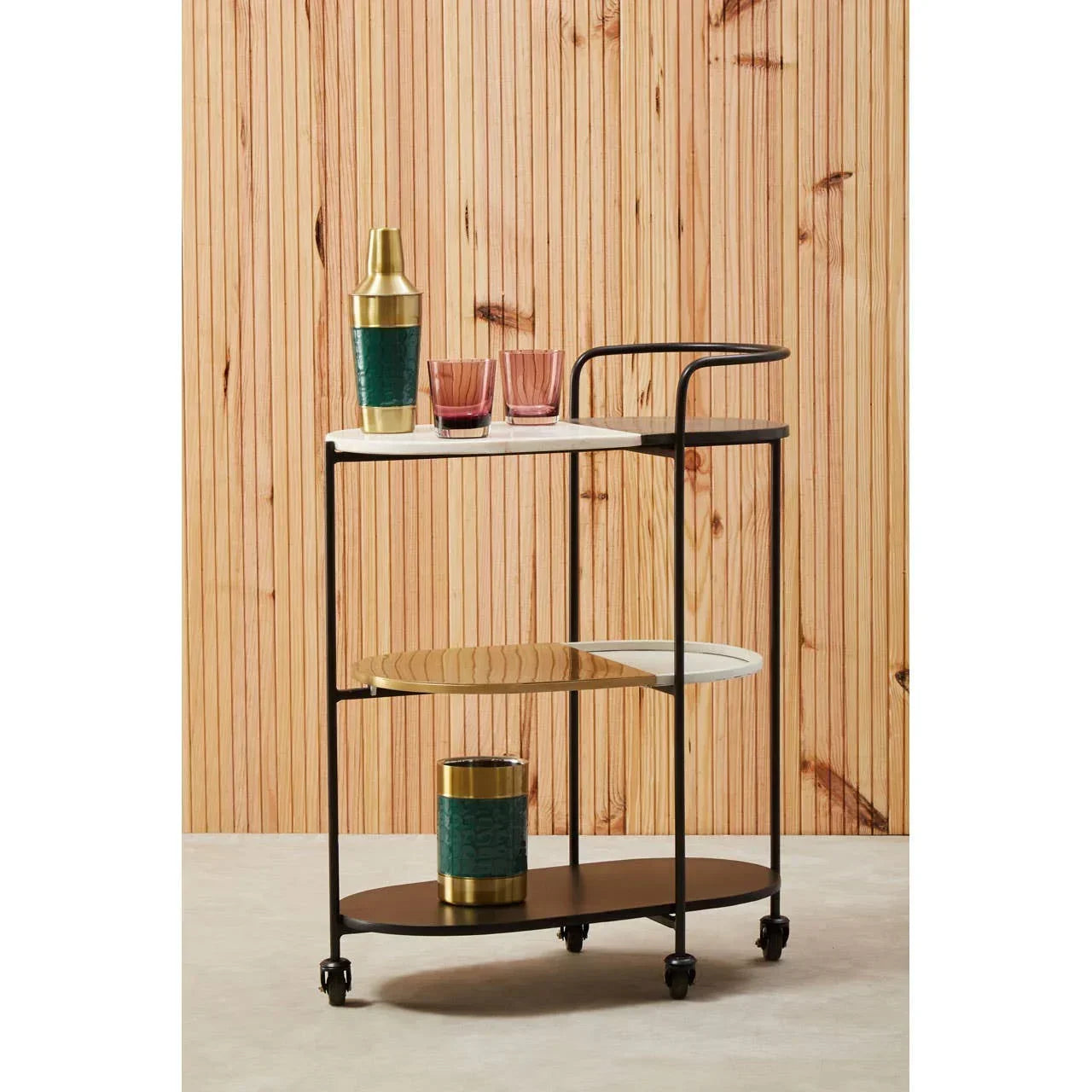 Three tier bar cart with marble effect shelves, gold cocktail shaker and glasses against wood panel wall