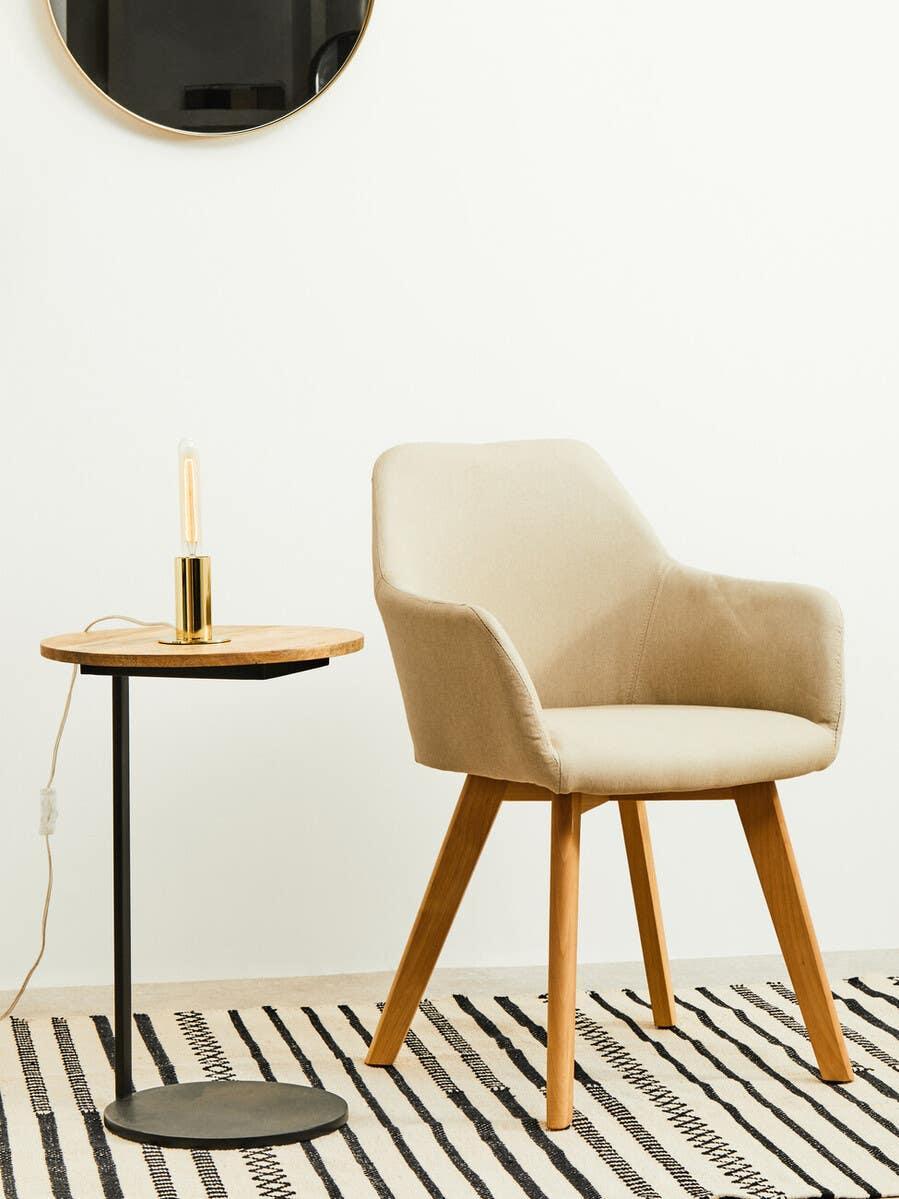 Beige fabric accent chair with wooden legs beside round side table and lamp on patterned rug