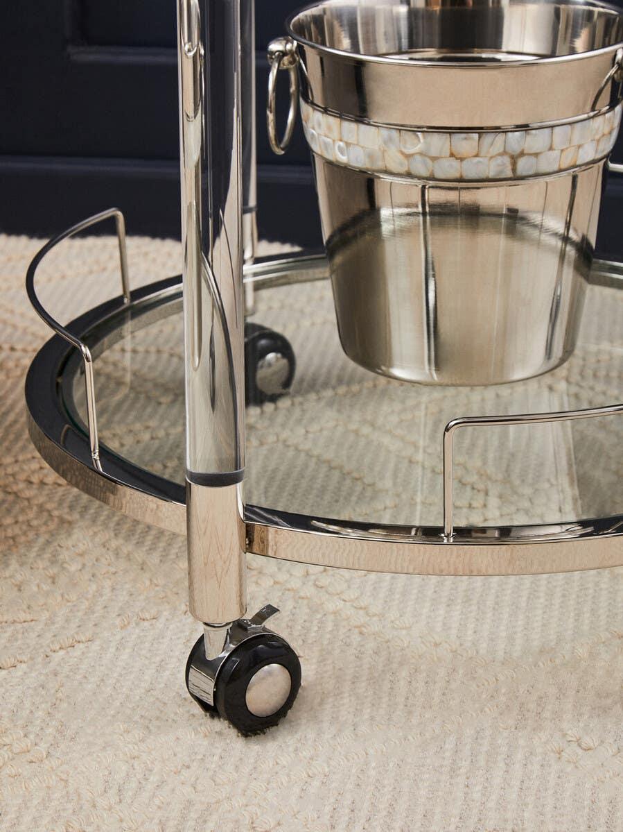 Chrome bar cart with glass shelf, metal ice bucket, and caster wheels on textured rug
