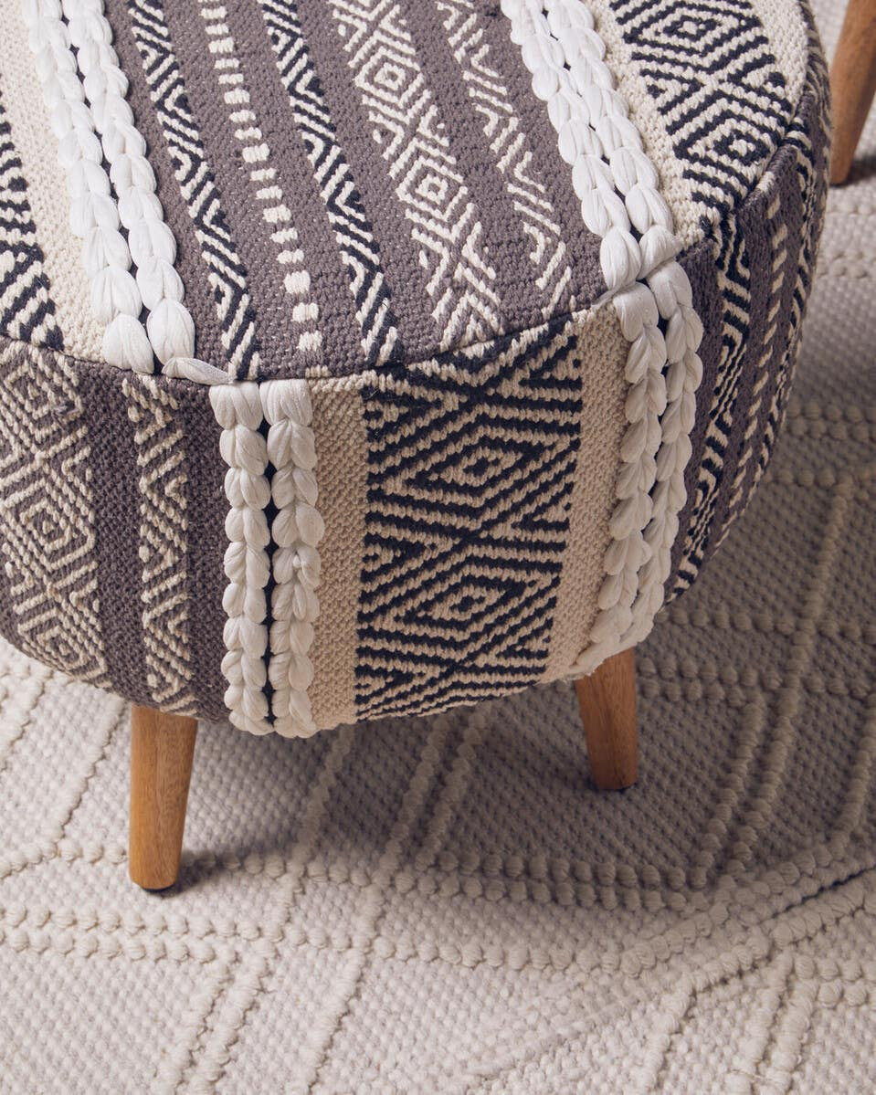 A grey and white patterned stool with a rounded top and wooden legs, placed on a textured light-colored rug. The fabric features various geometric designs in shades of grey, cream, and white