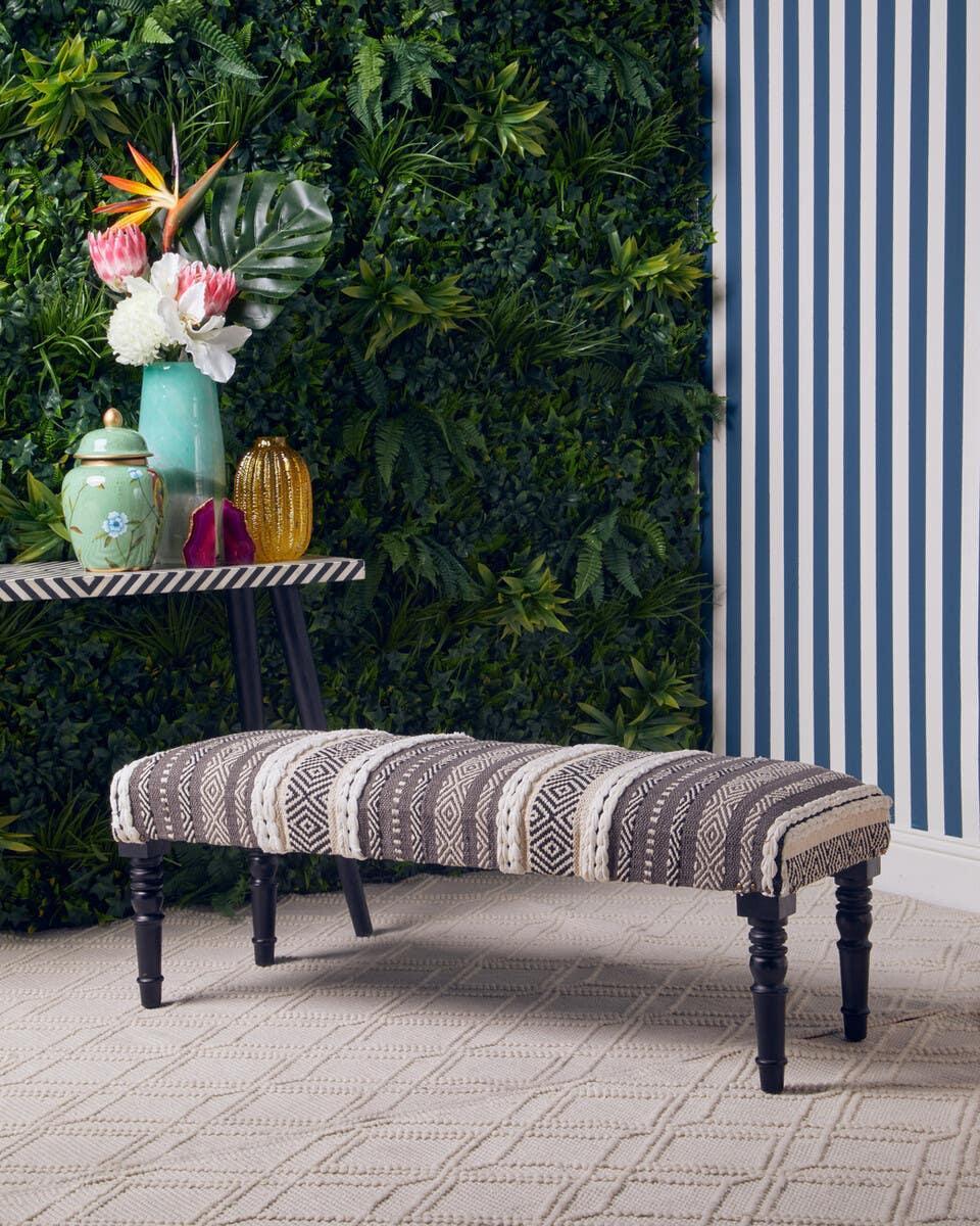 Patterned upholstered bench with dark wooden legs on textured rug beside feature table