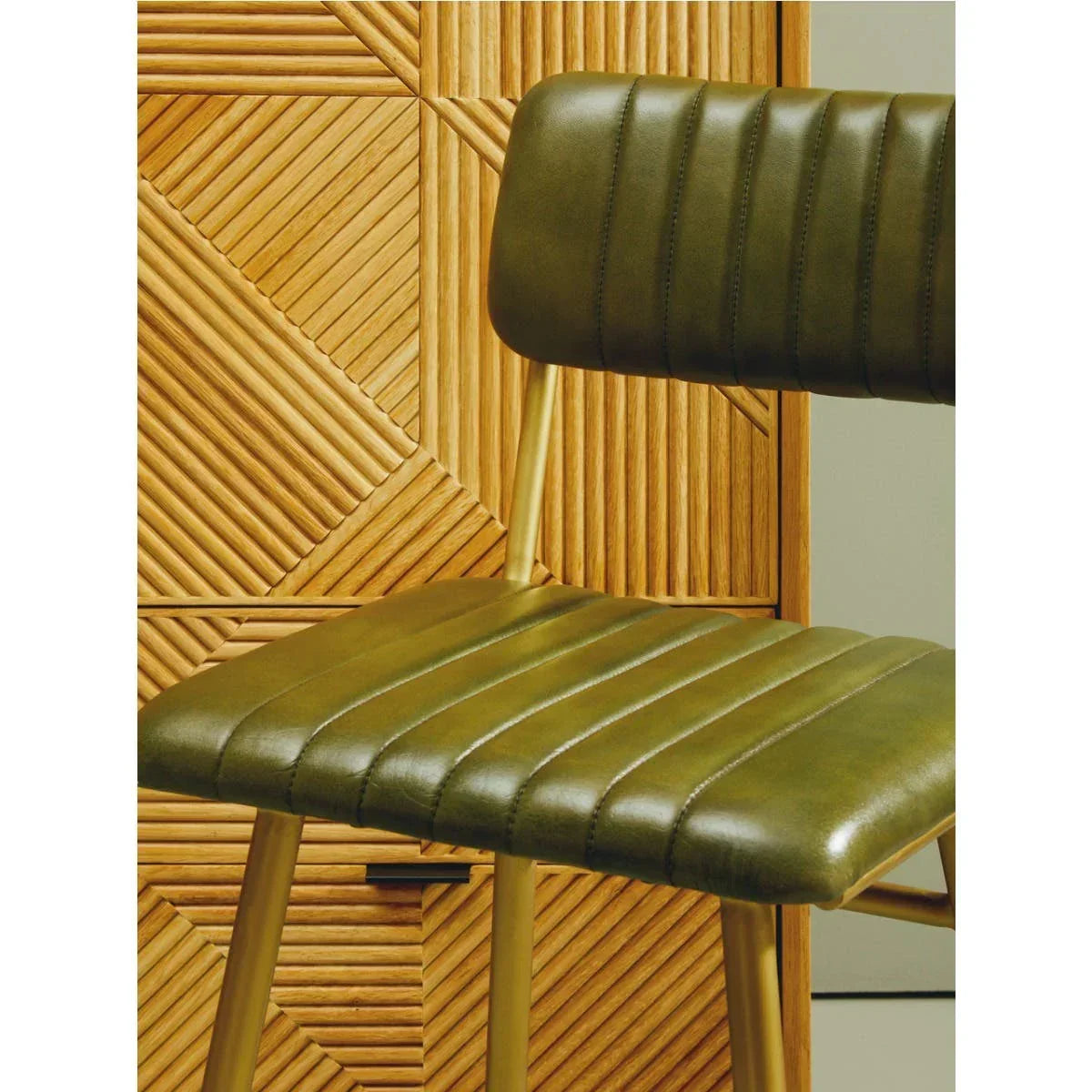 Olive green faux leather dining chair with stitched detail and gold legs beside wood cabinet