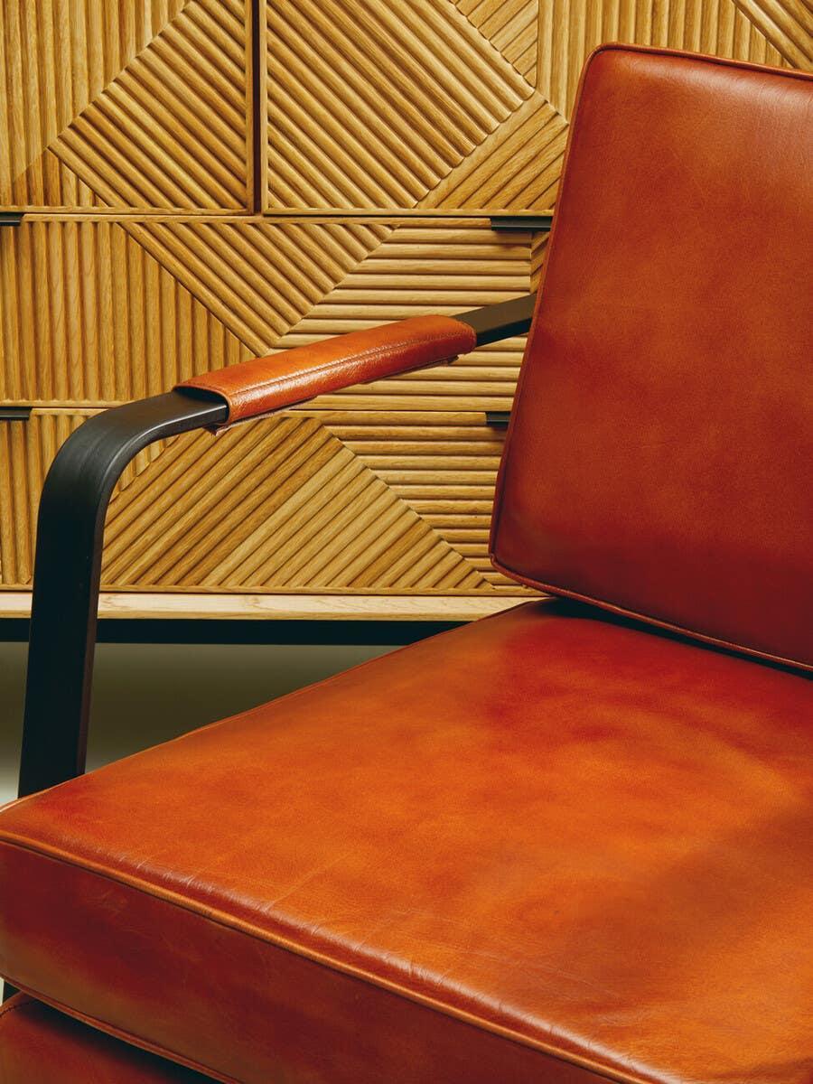 Brown leather armchair with black metal armrest in front of geometric wood paneling
