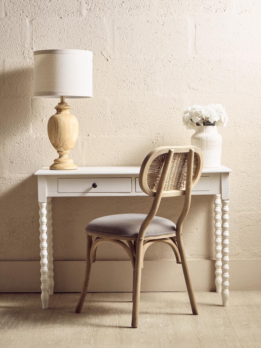 White wooden desk with spindle legs, rattan chair, table lamp and ceramic vase with flowers