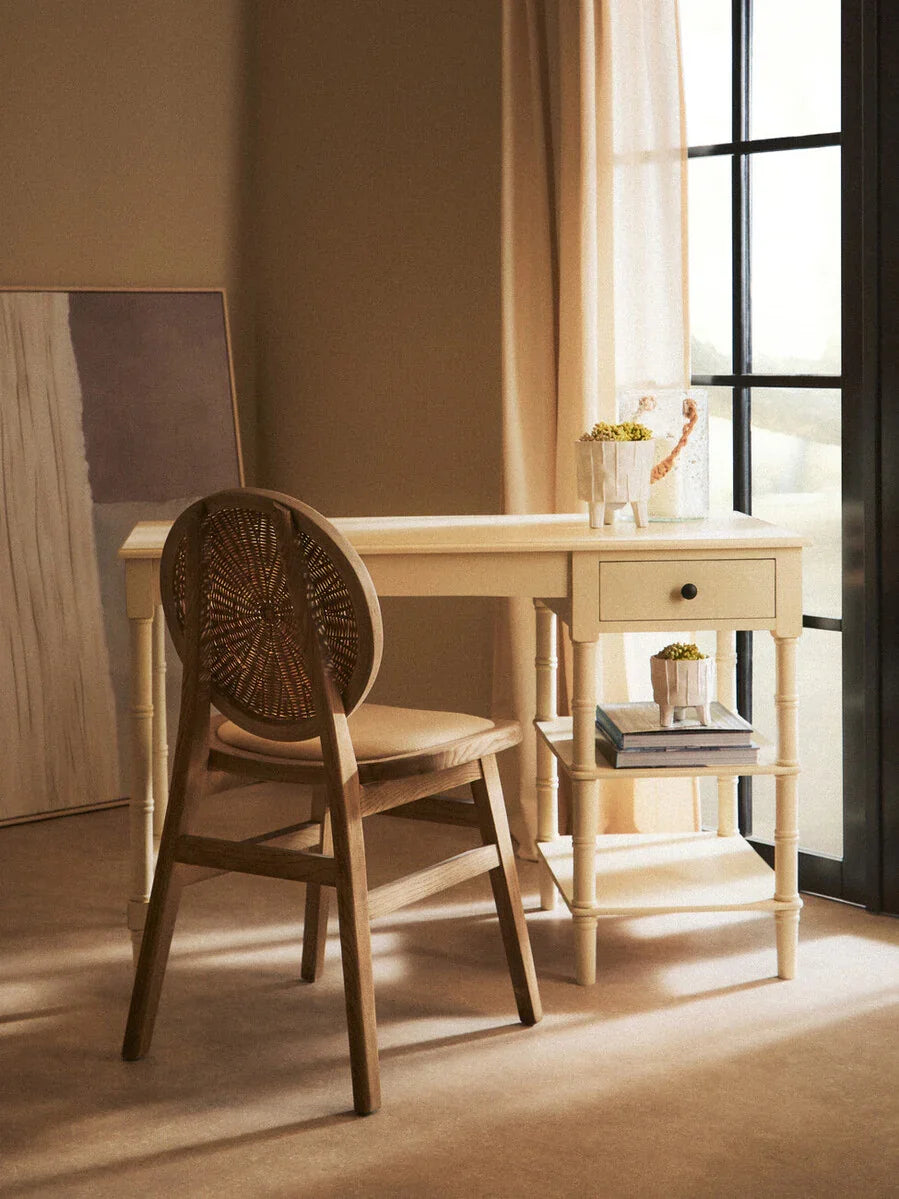 Wooden desk with drawer and shelf beside window, paired with rattan back chair