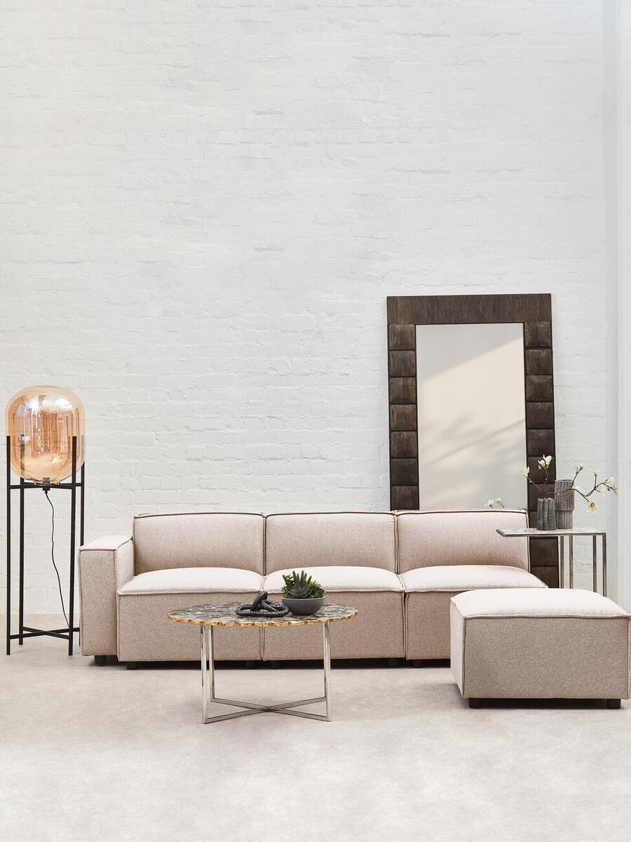 A light beige armless modular sofa with plush cushions is positioned against a neutral wall. The sofa features clean lines and a contemporary design. Next to it, a matching ottoman complements the seating arrangement. A tall mirror with a dark frame leans against the wall, reflecting the room's light. A decorative plant sits on a sleek, modern side table nearby, adding a touch of greenery to the space