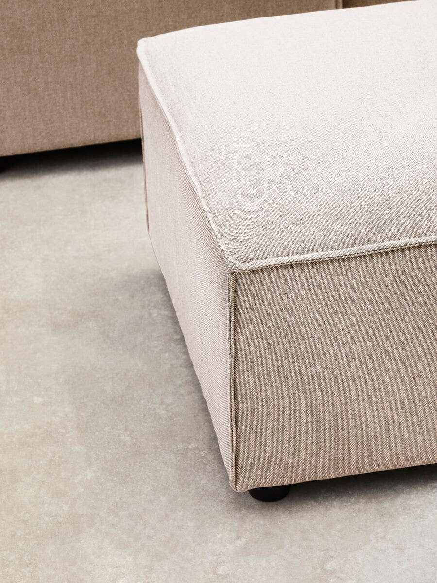 A corner view of a modular ottoman upholstered in natural fabric, featuring a soft, square top and clean lines. The ottoman has a light beige color with a subtle texture, resting on a smooth surface