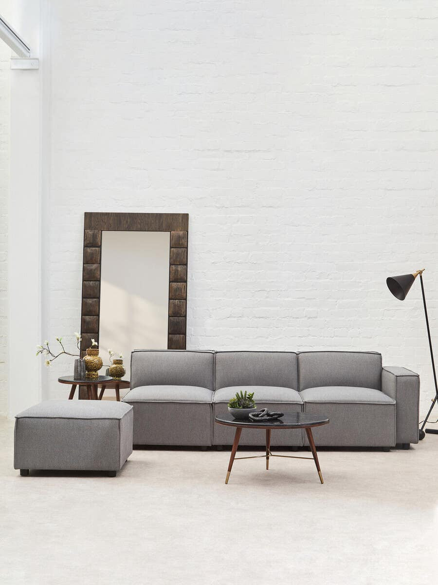 Grey fabric right arm modular sofa with plush cushions, accompanied by a matching ottoman, set against a light-colored wall. A large mirror and a floor lamp are visible in the background, adding to the modern decor