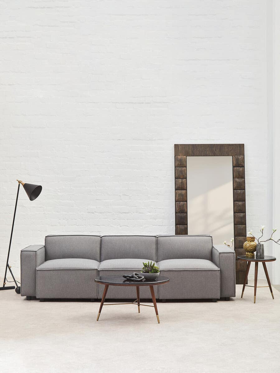 A grey fabric armless modular sofa with three cushions, positioned in a bright room featuring a large mirror and a floor lamp. A small potted plant and a side table are visible next to the sofa, with a light-colored floor and white walls in the background