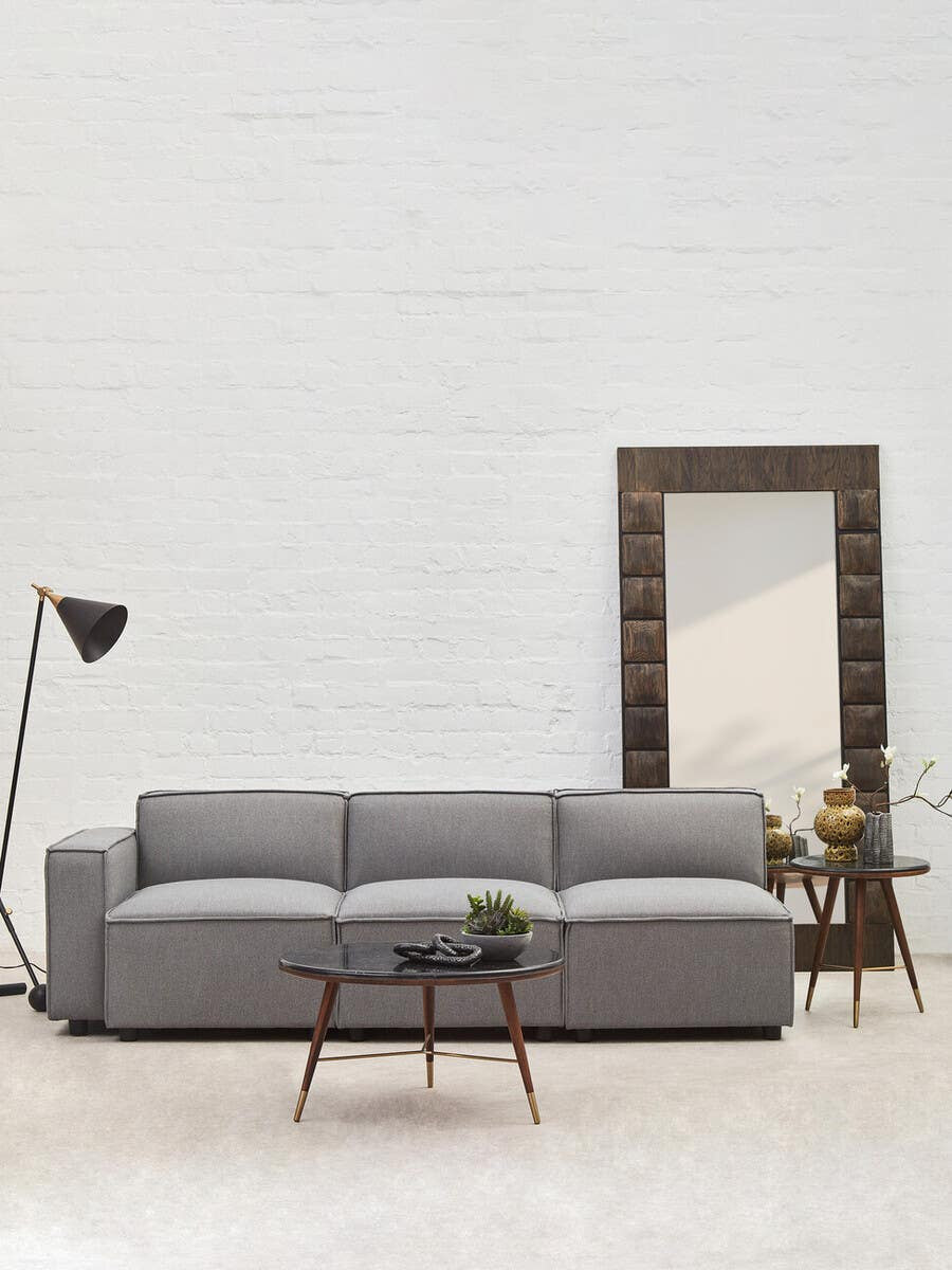 Grey fabric armless modular sofa with three plush cushions, set against a light-colored wall. A tall floor lamp stands to the left, and a decorative mirror with a wooden frame is positioned behind the sofa. A small potted plant is placed on a side table next to the sofa