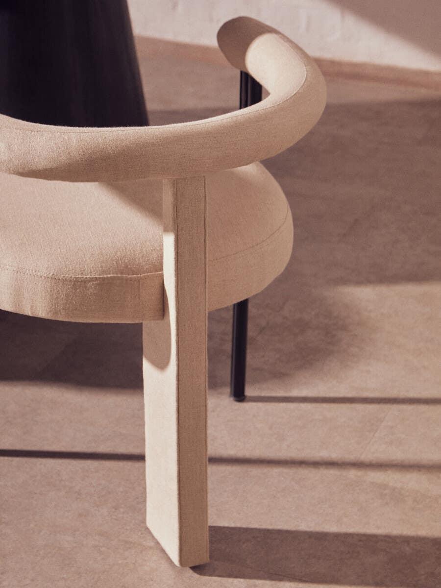 Curved beige upholstered armchair with fabric legs and rounded backrest on tile floor