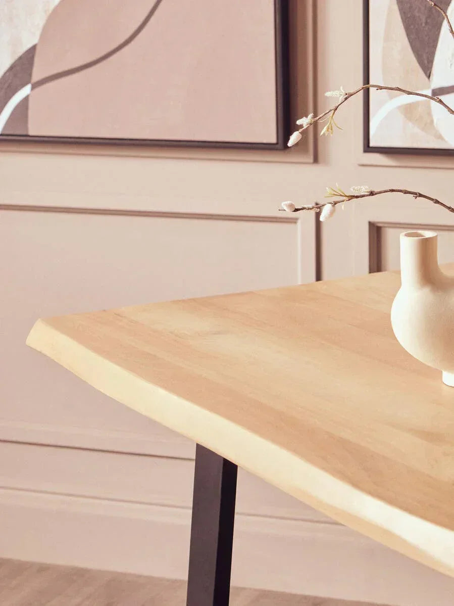 Light wood dining table with black metal leg, ceramic vase and wall art in background