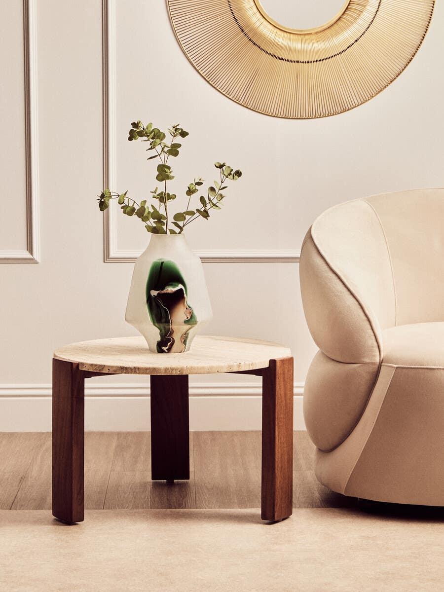 Round wooden side table with marble top and decorative vase beside cream accent chair