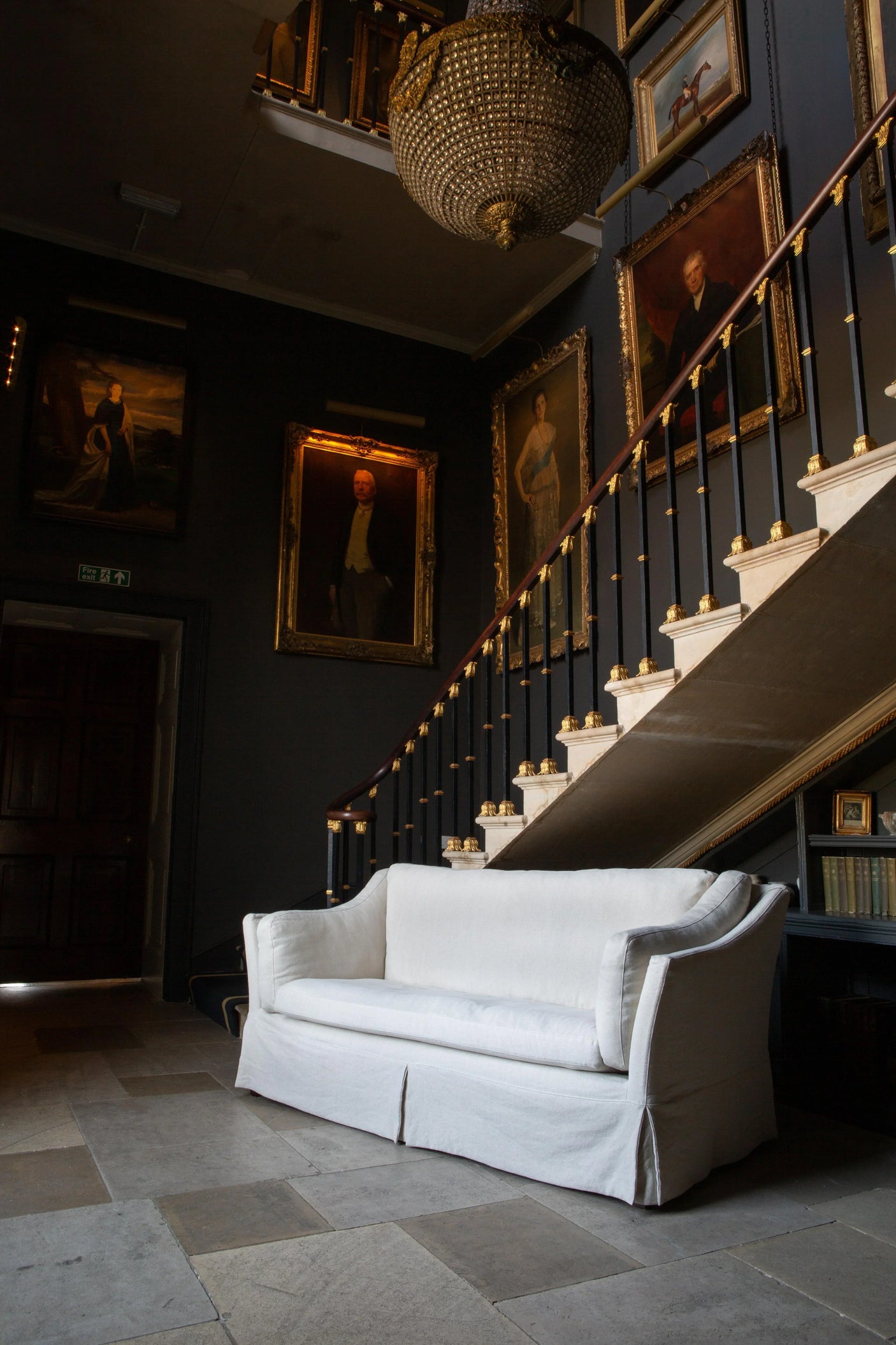 White fabric sofa placed in a classic hallway beneath staircase and framed portraits
