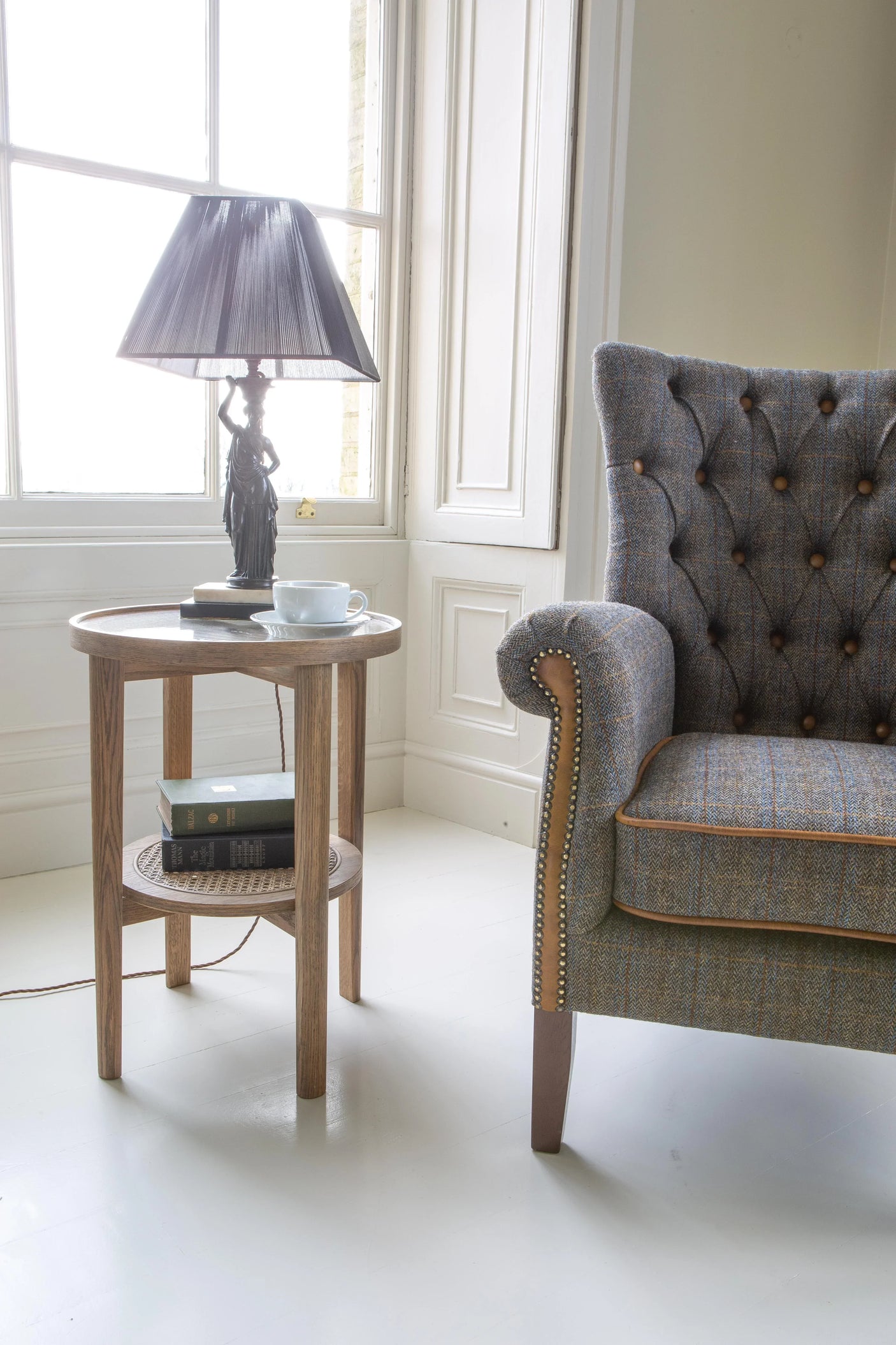 Upholstered armchair beside round wooden side table with lamp, books and teacup