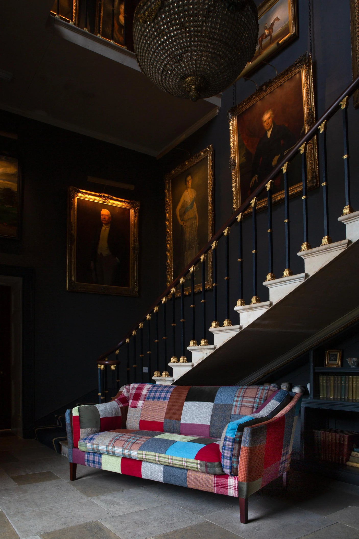 Patchwork fabric sofa in a classic hallway with dark walls, gold-framed portraits, and staircase