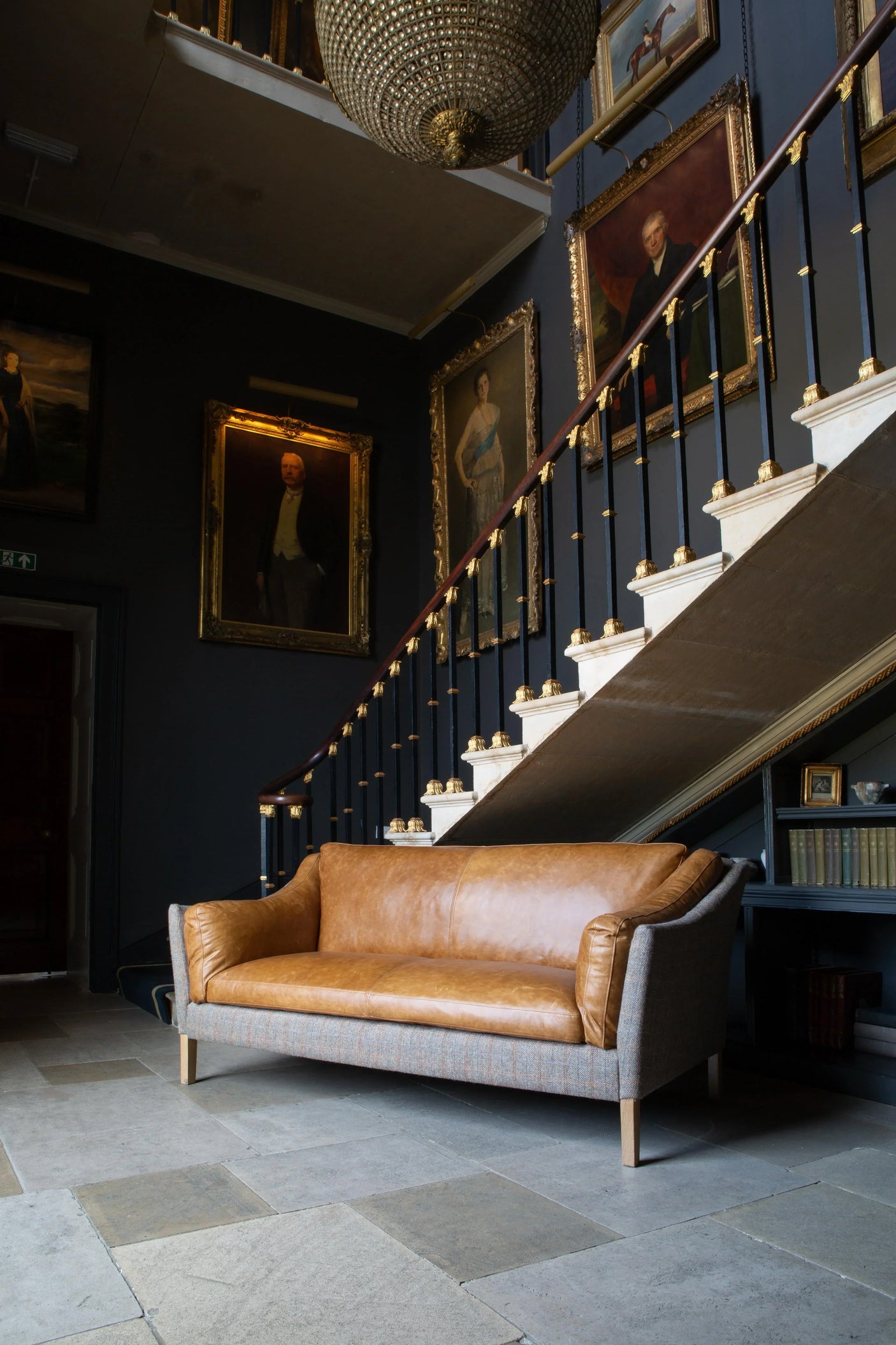 Tan leather and fabric sofa in front of staircase with framed portraits and chandelier