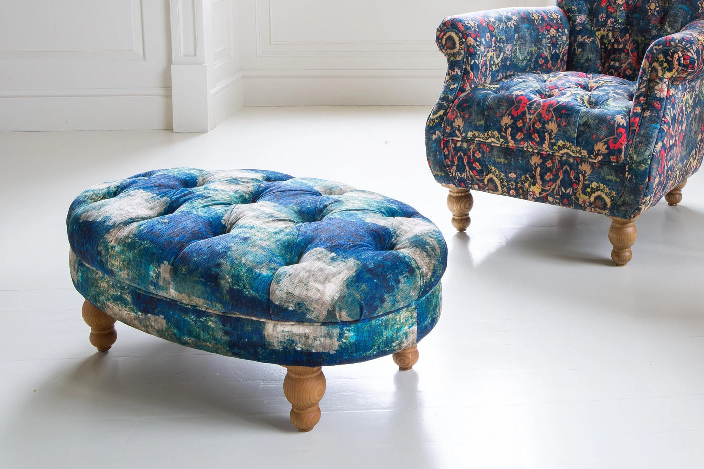 Blue velvet ottoman with button tufting and wooden legs beside patterned armchair