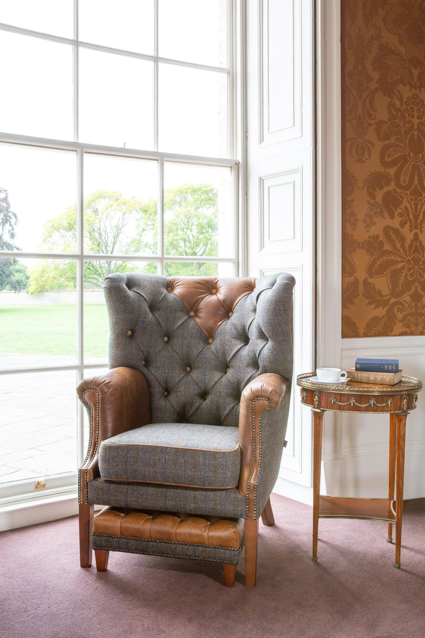 Leather and fabric accent armchair with matching footstool near window and antique side table