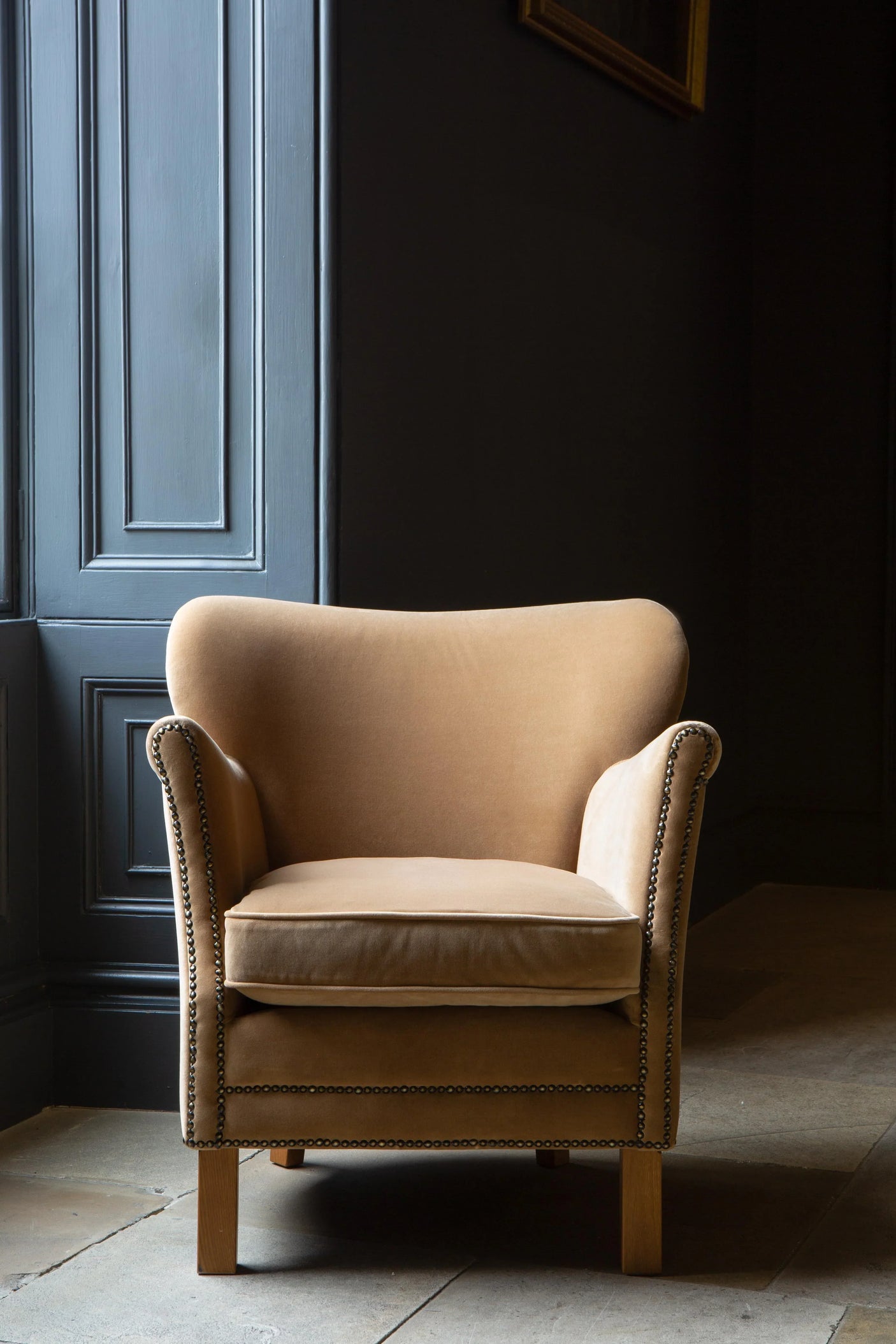 Beige velvet accent chair with wooden legs and studded trim beside dark panelled wall