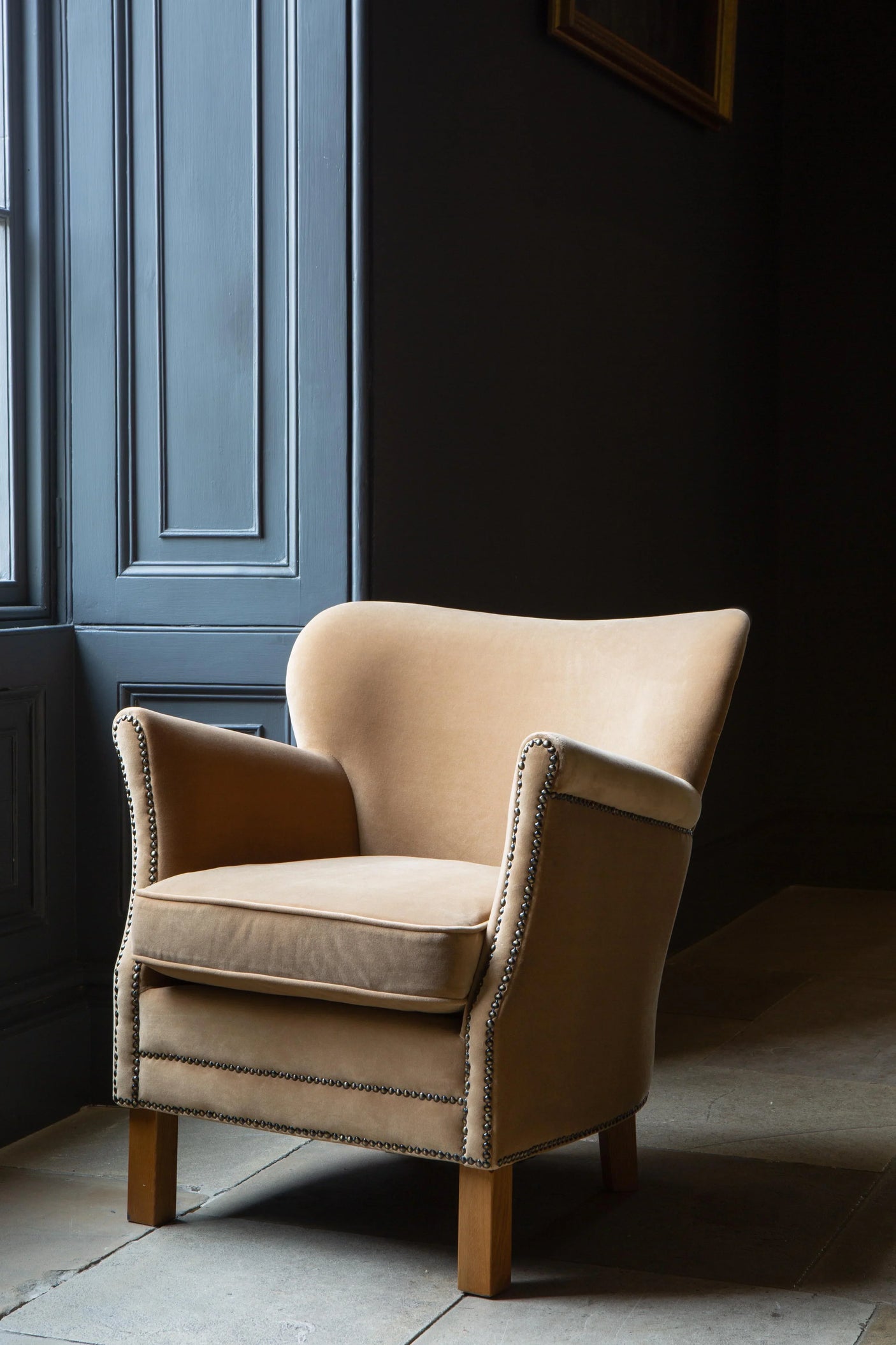 Beige velvet accent armchair with nailhead trim and wooden legs next to blue panelled wall