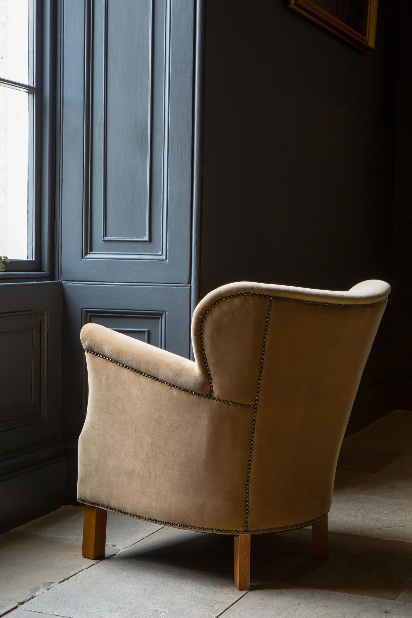 Tan velvet armchair with studded trim and wooden legs beside a window in a modern room