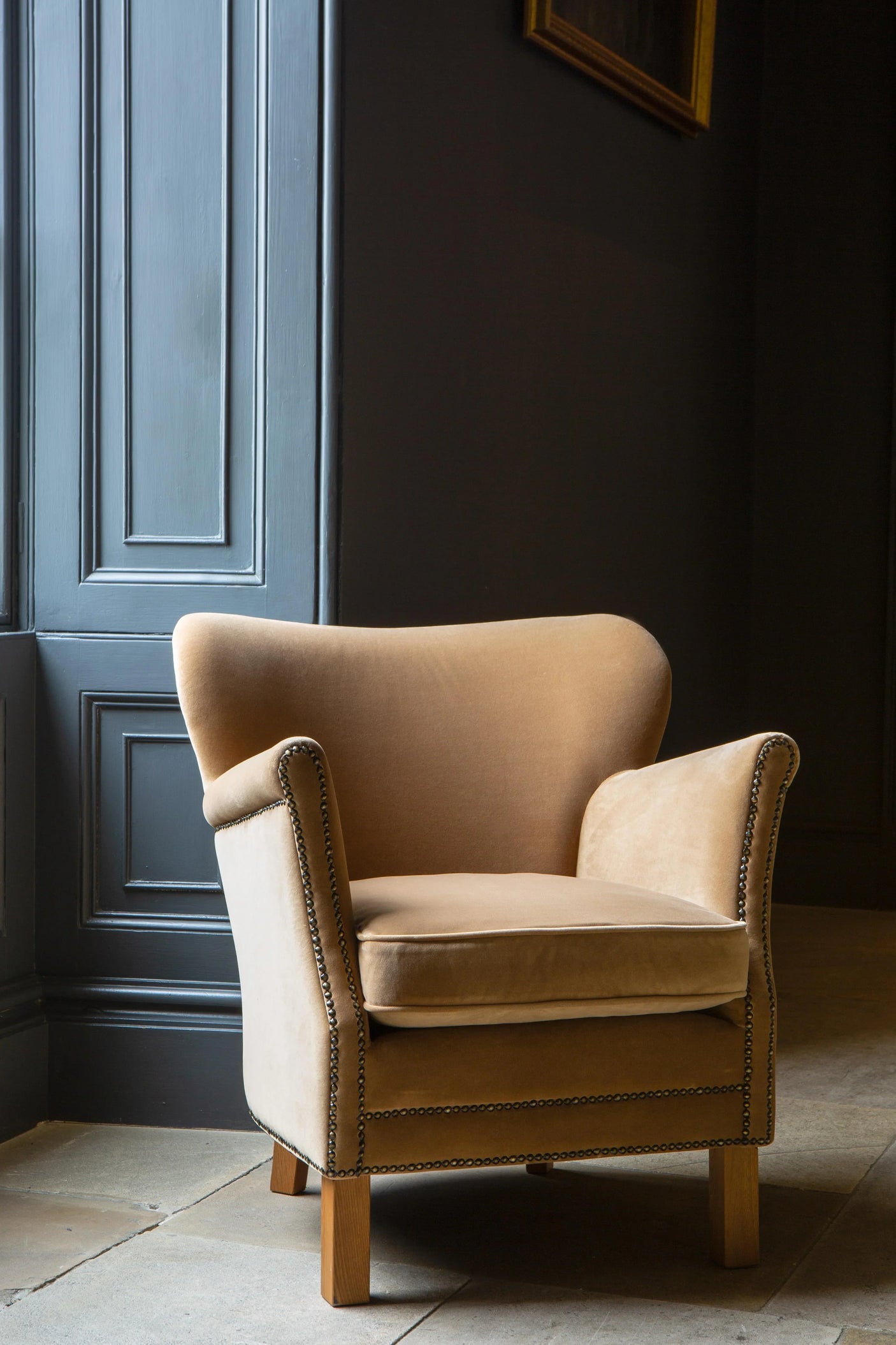 Beige fabric armchair with studded trim and wooden legs beside a window