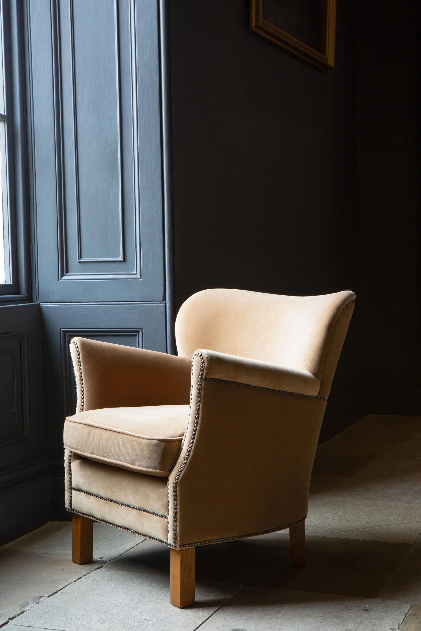 Tan fabric armchair with nailhead trim and wooden legs by a window