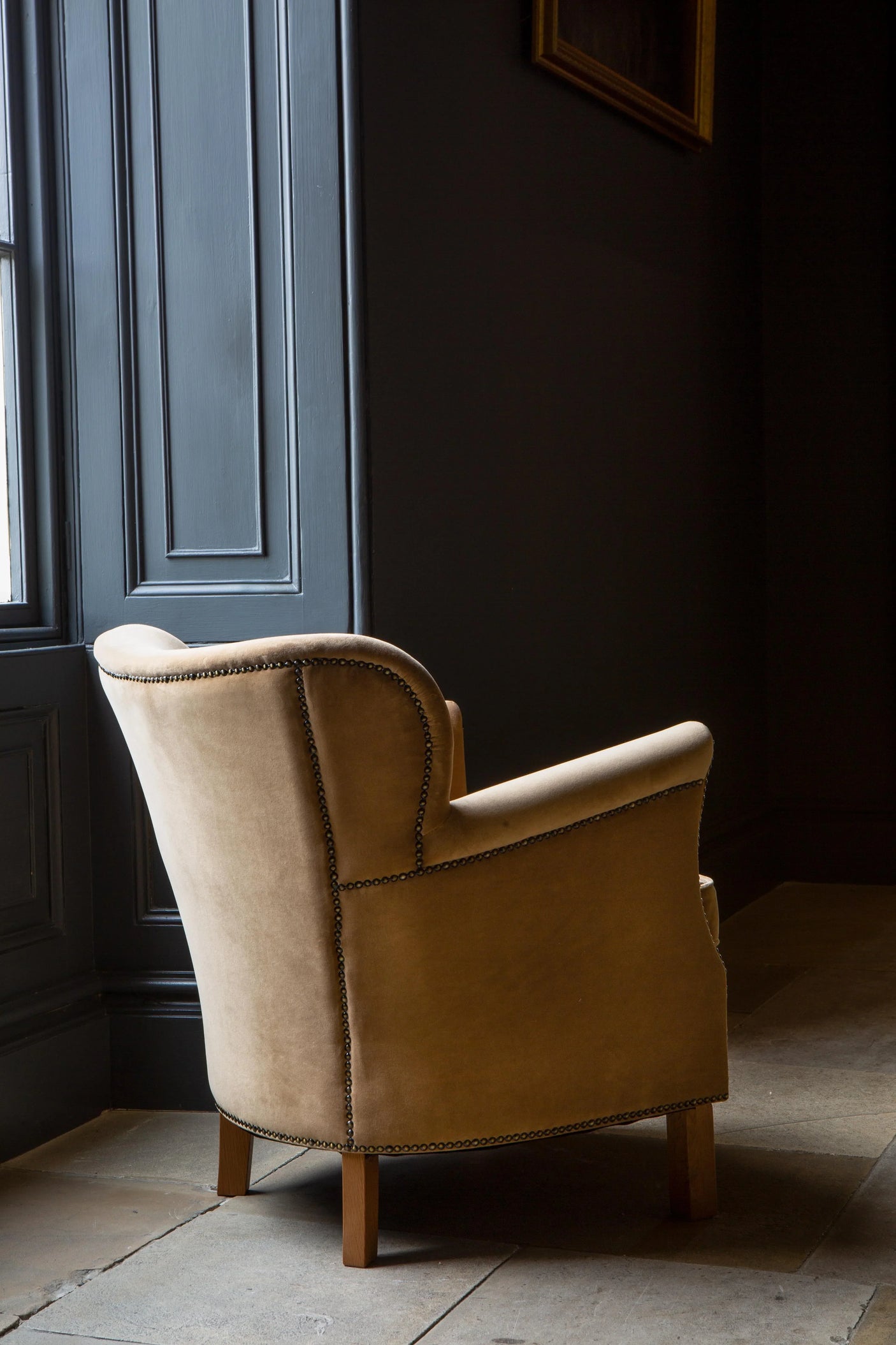Tan fabric armchair with studded trim and wooden legs beside window on stone floor