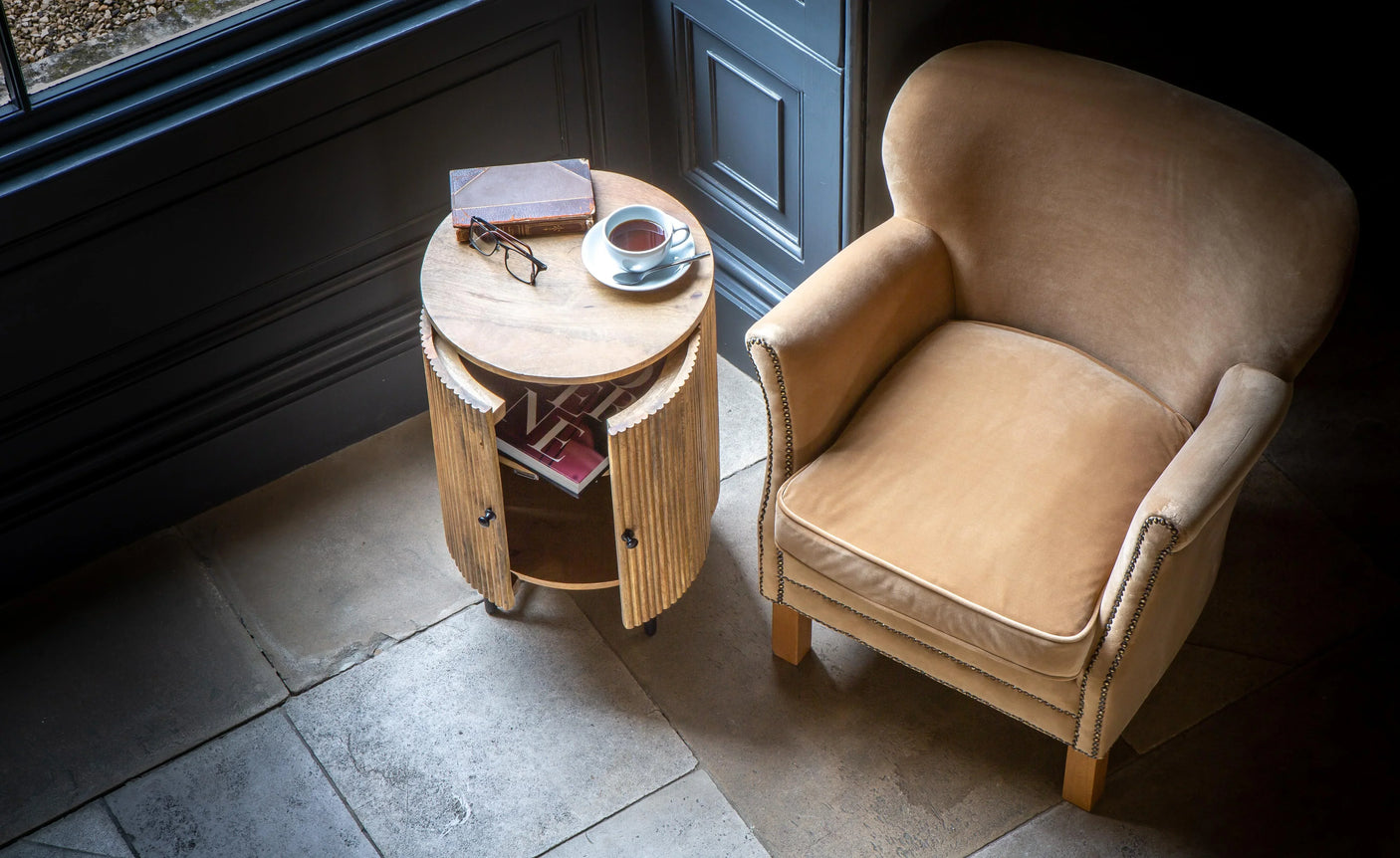 Tan velvet armchair beside round wooden side table with open storage and cup of tea