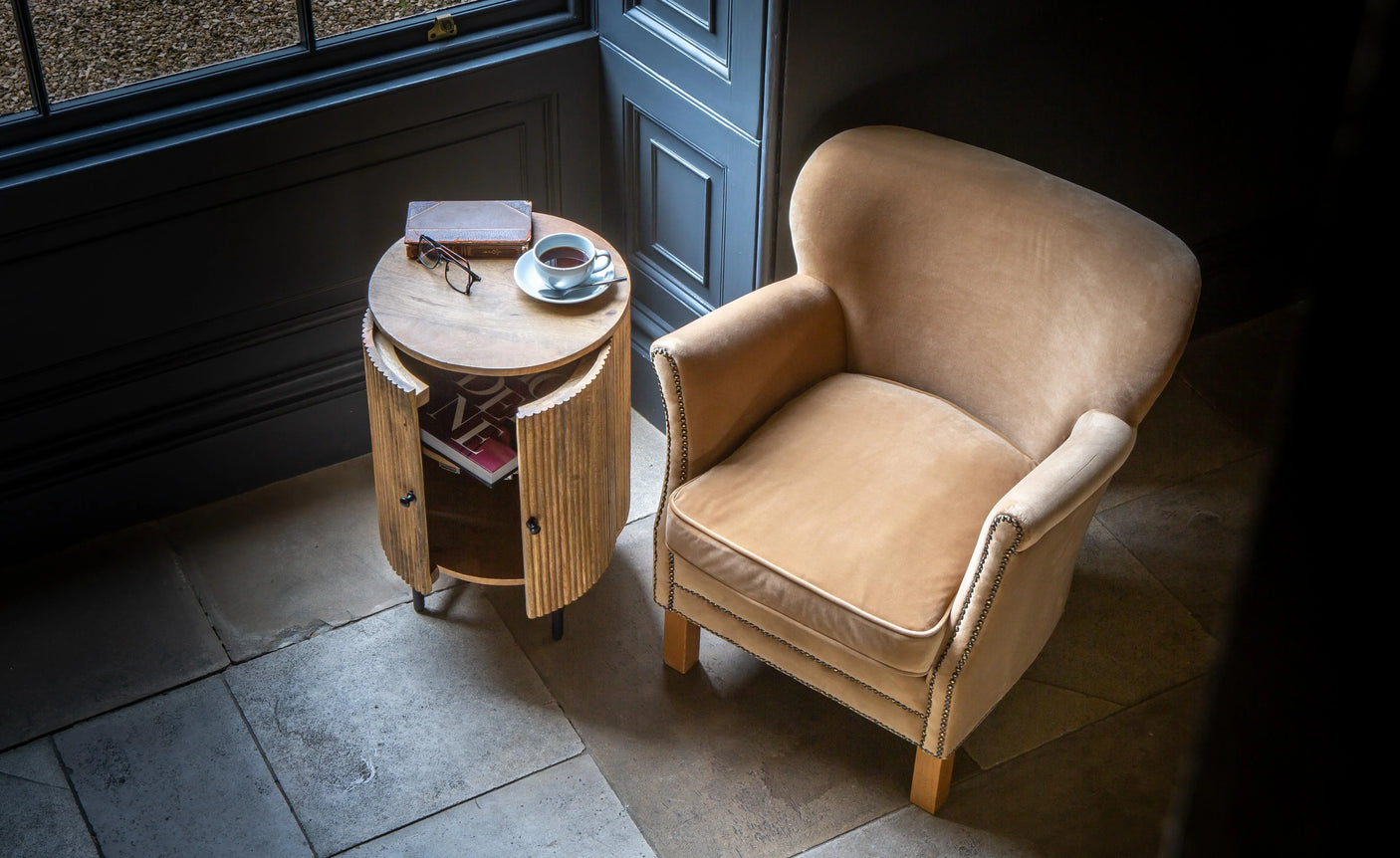 Tan fabric armchair paired with round wooden side table holding book and cup in corner room