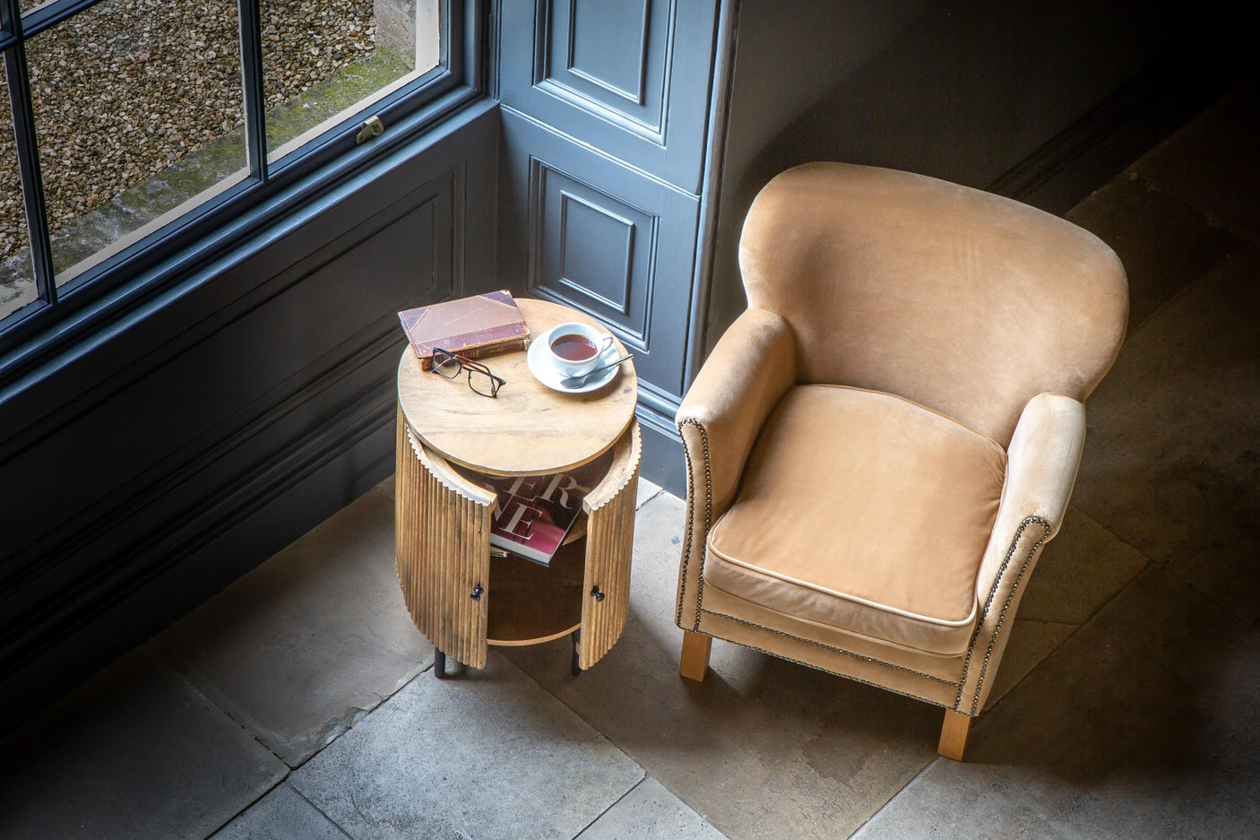 Beige armchair beside round wooden side table with cup of tea and book by window
