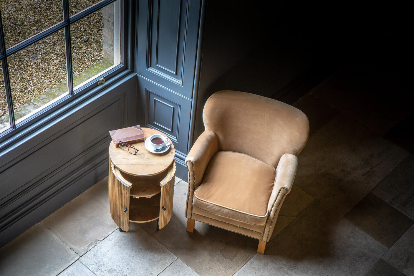 Beige velvet armchair beside wooden side table with book, glasses, and cup in a window corner