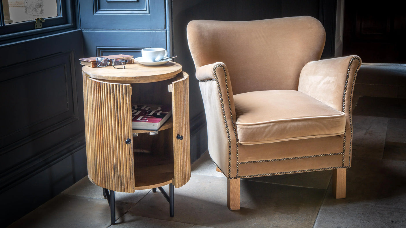 Velvet accent armchair beside round wooden side table with open door and storage shelf