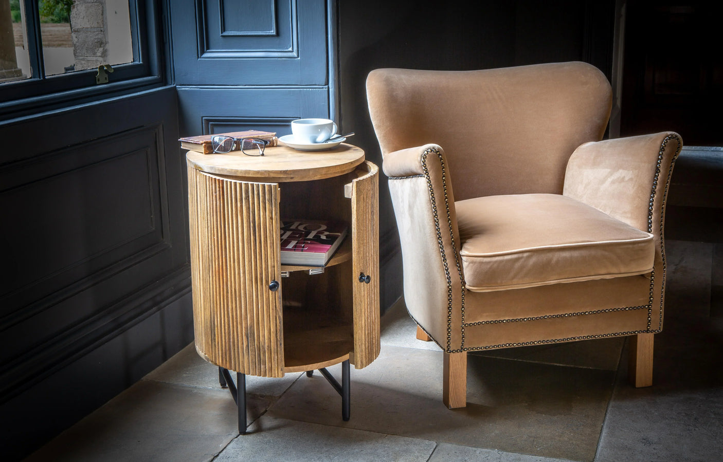 Wooden side table with open slatted doors beside beige velvet armchair