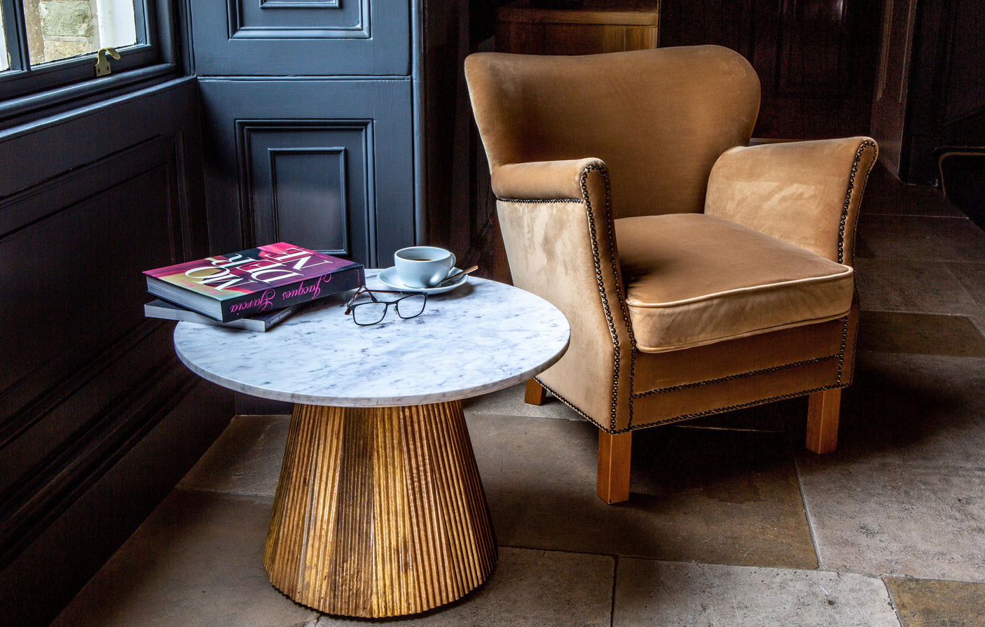 Velvet armchair beside marble top coffee table with books, cup and glasses