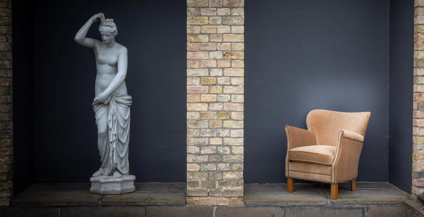 Beige upholstered armchair with wooden legs beside stone statue against brick and dark wall