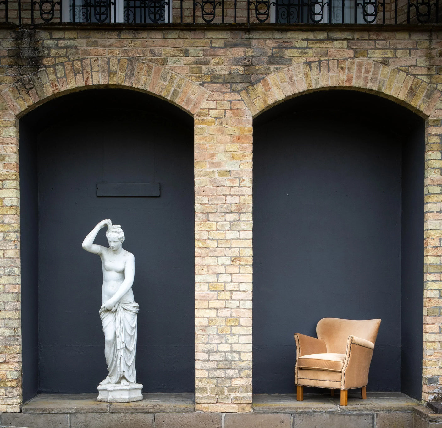 Modern tan accent armchair with wooden legs beside a classical stone statue under brick arches