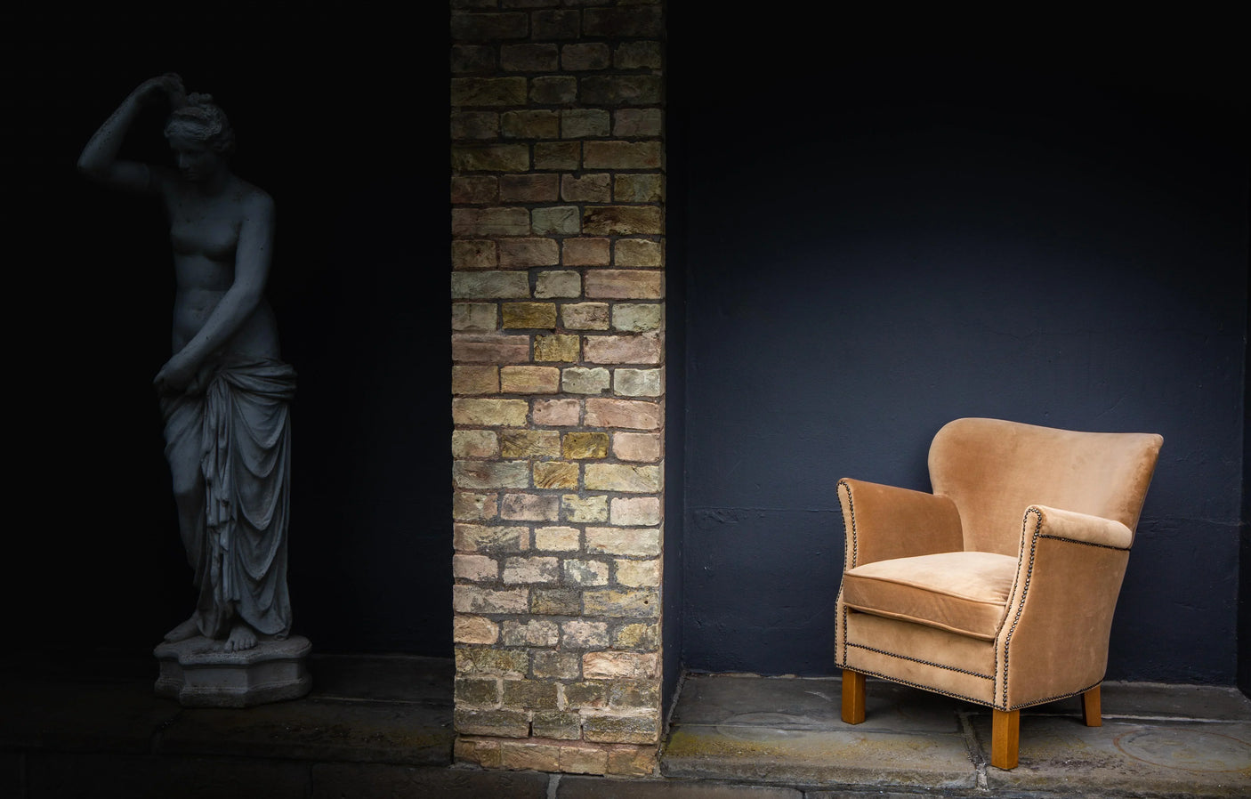 Beige velvet armchair with studded trim beside brick wall and classic statue