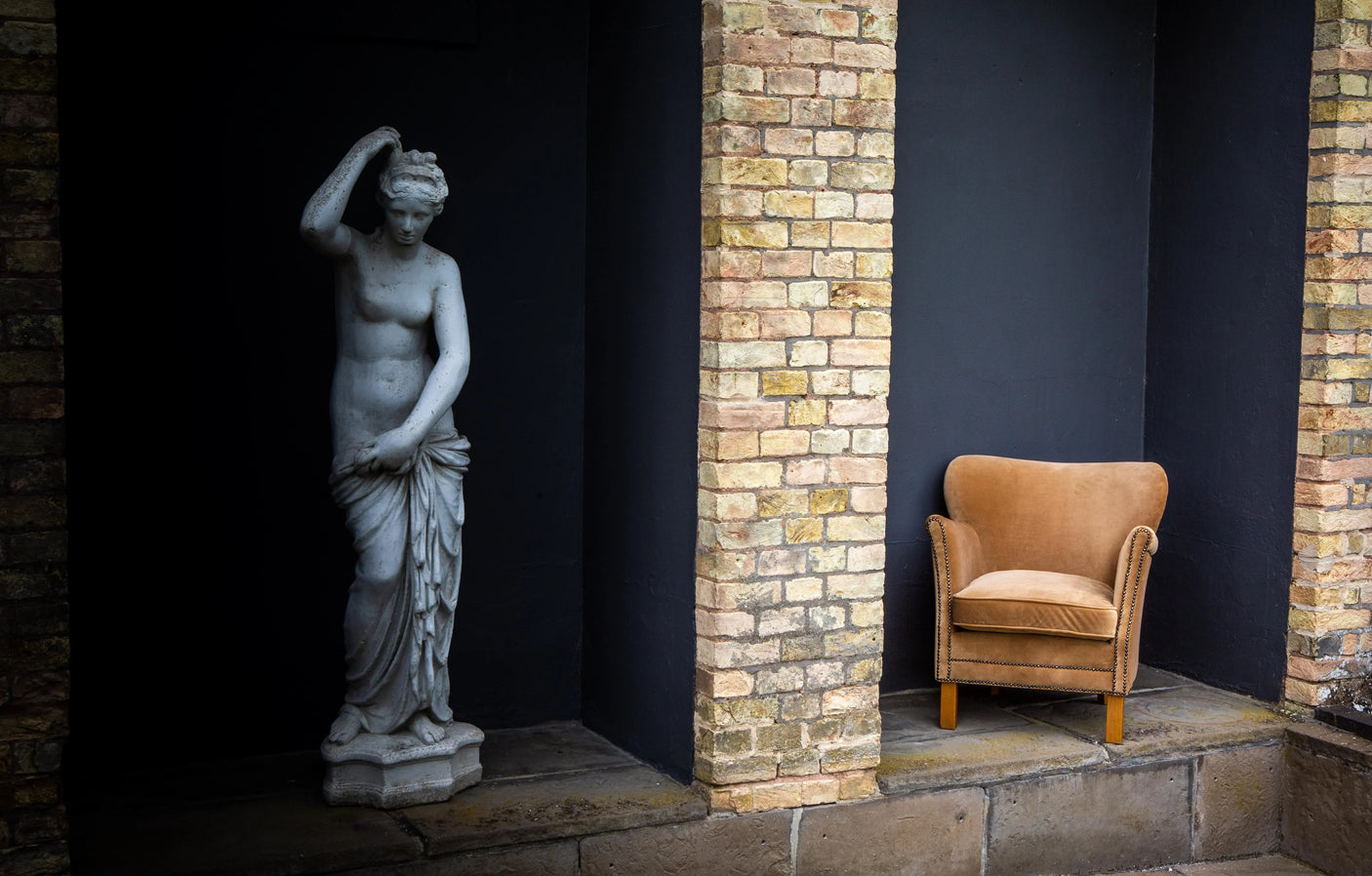 Tan velvet accent chair with nailhead trim beside classical stone statue and brick pillars