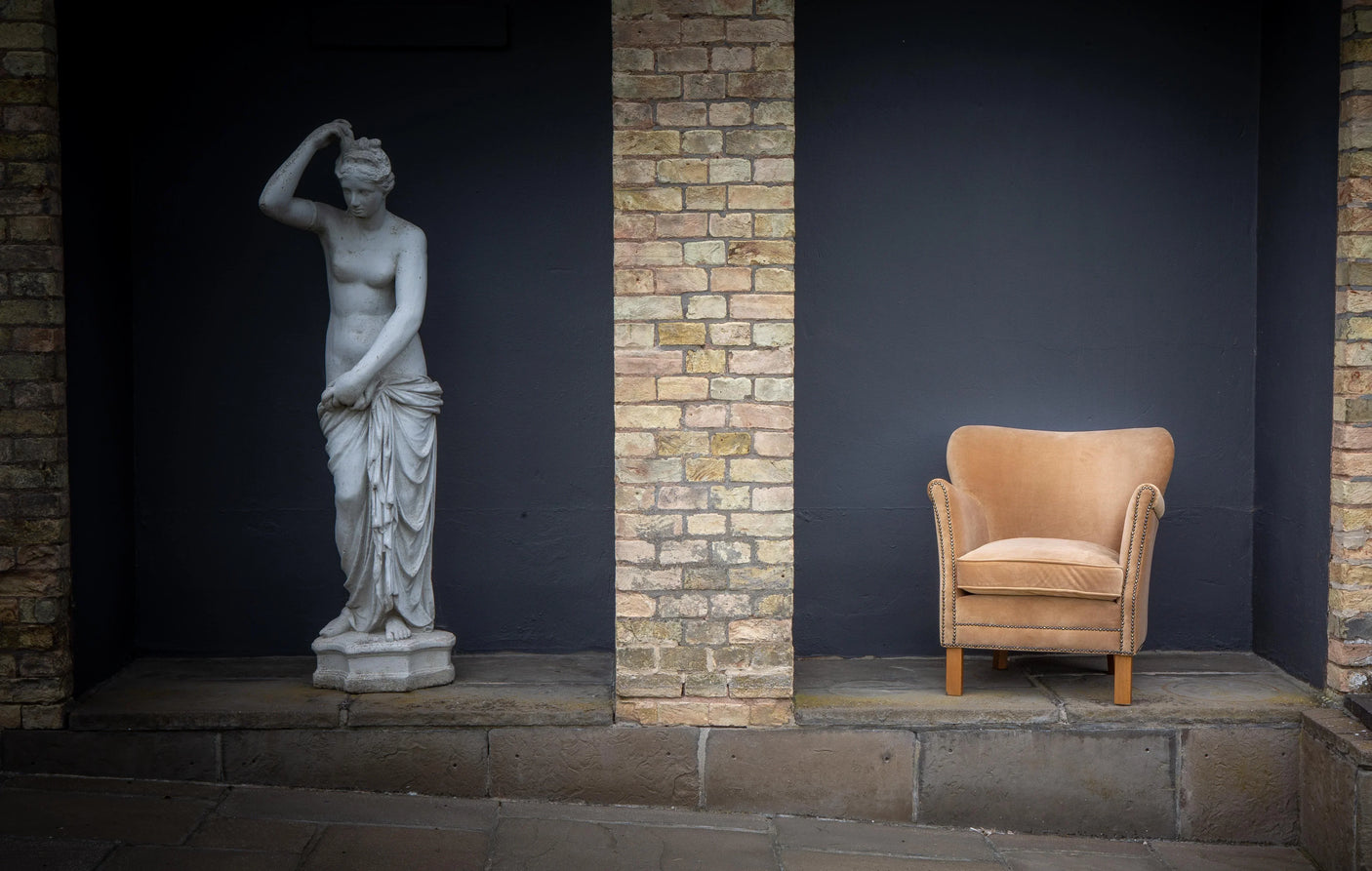 Beige velvet armchair with wooden legs beside classical statue, brick wall background