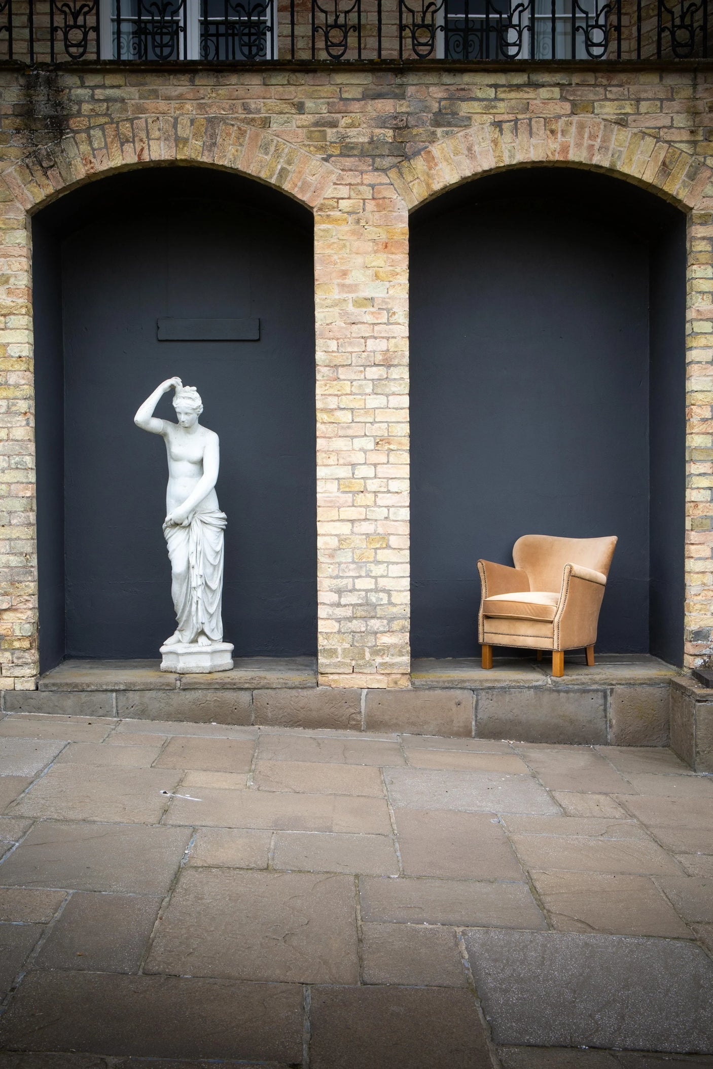 Tan upholstered armchair with wooden legs beside classical statue against brick wall