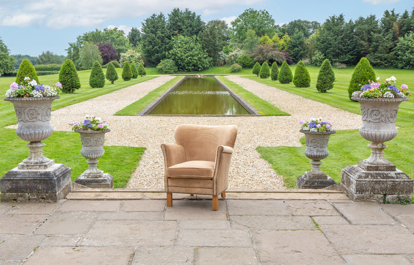 Beige fabric armchair with wooden legs in a formal garden setting surrounded by stone planters