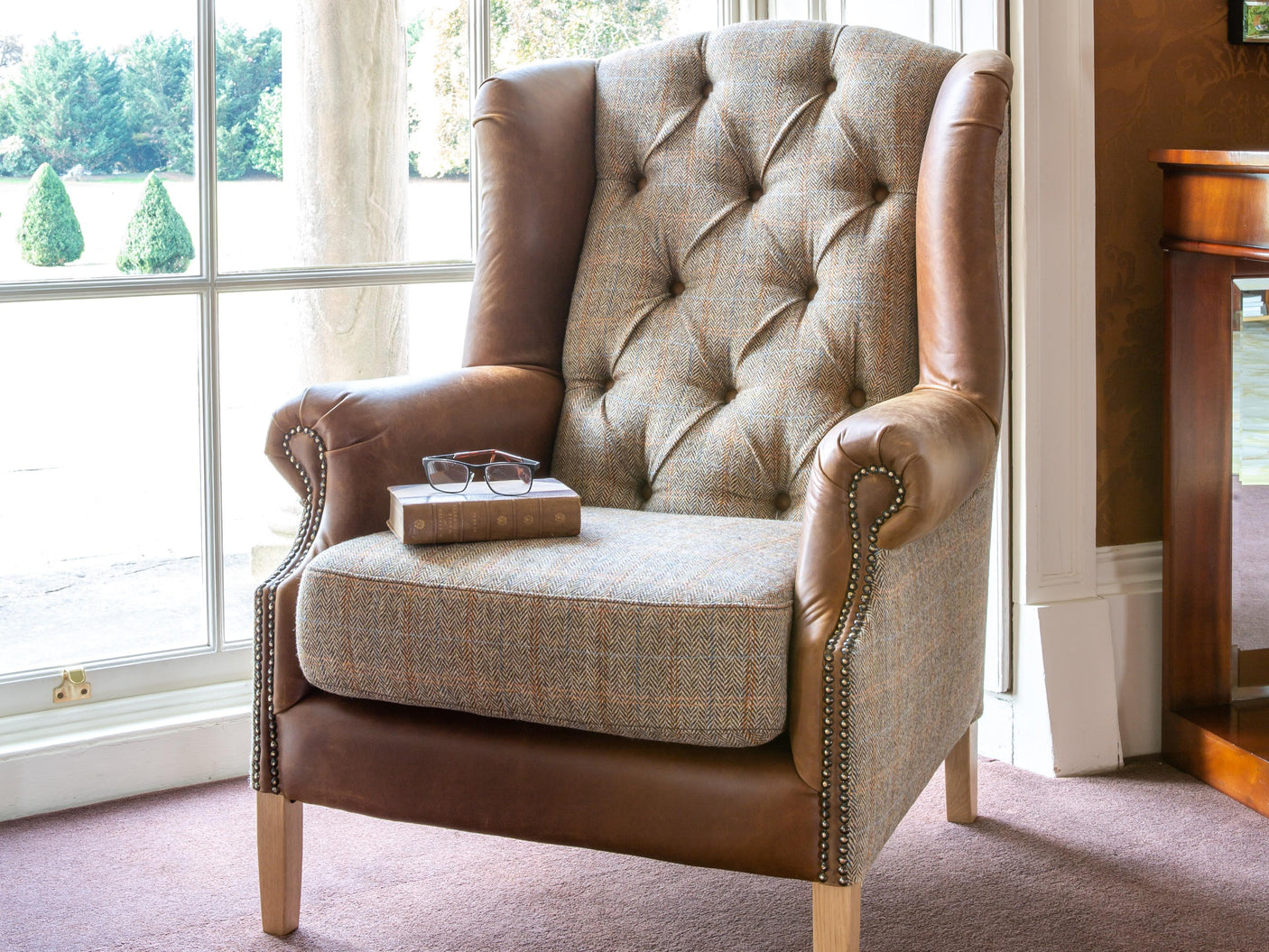 Button-tufted wingback armchair with tweed fabric and brown leather accents by window