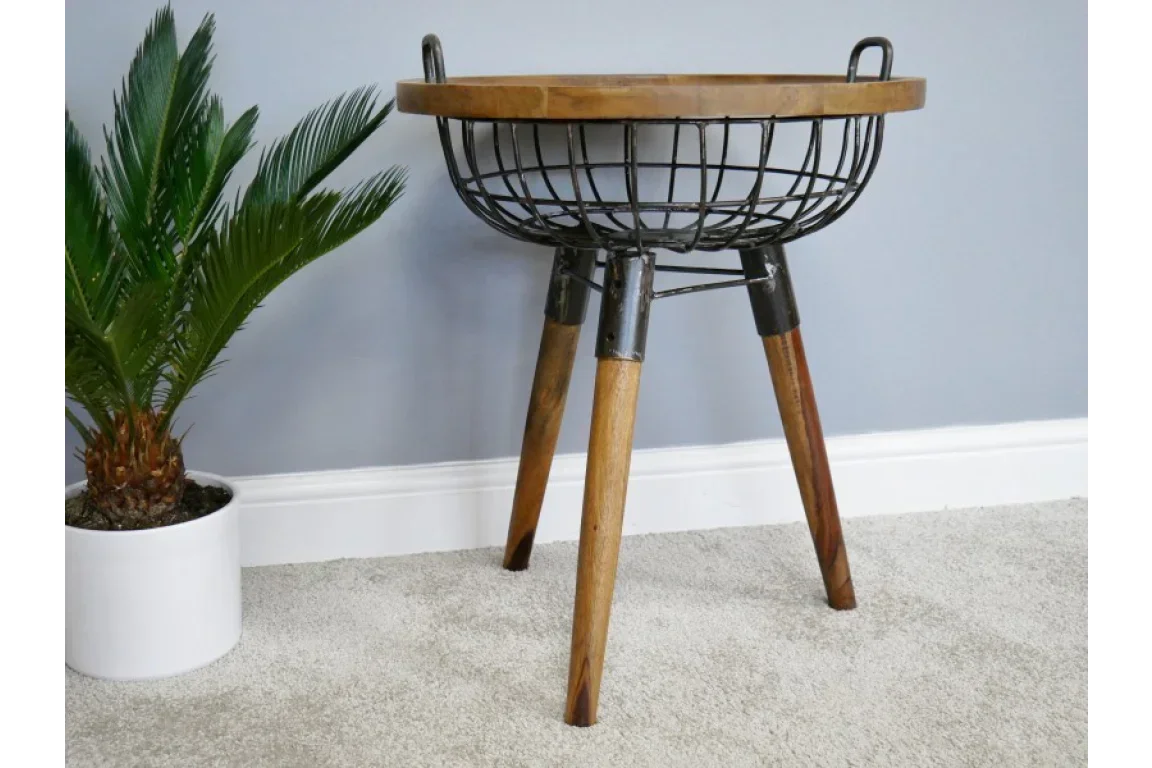 Industrial side table with wire basket base and wooden top beside potted plant on carpet