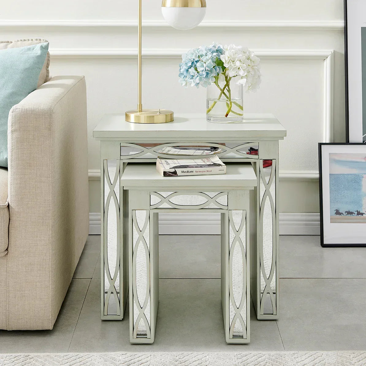 Mirrored nesting tables beside sofa with lamp, books, and floral arrangement