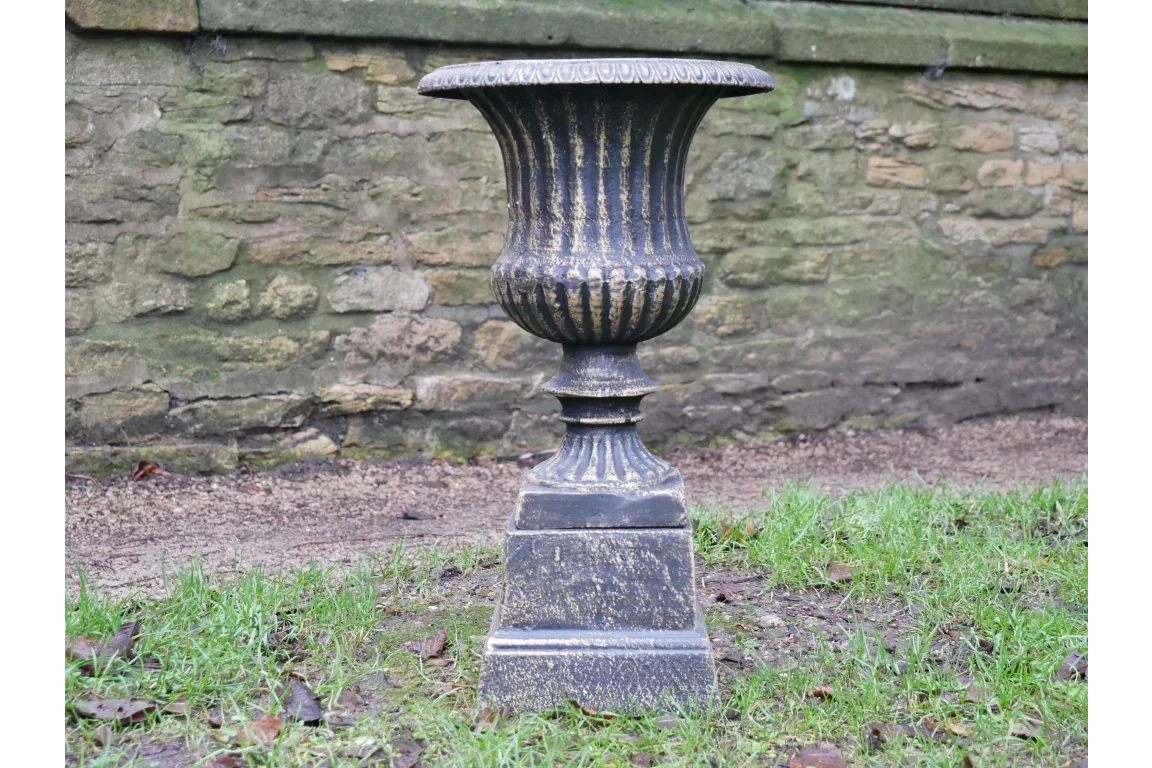 Black metal garden planter urn with fluted detailing on grass by stone wall