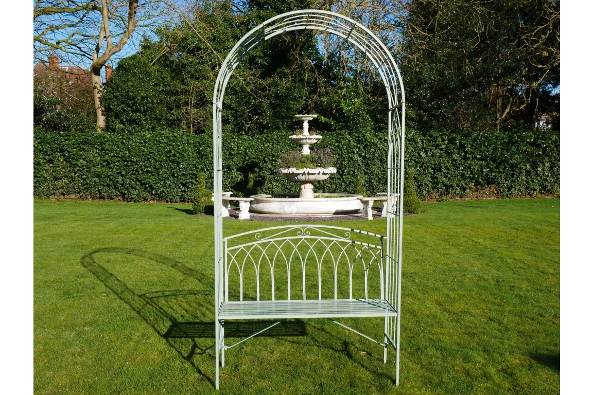 Green metal garden bench with arched trellis, placed on lawn near a stone fountain