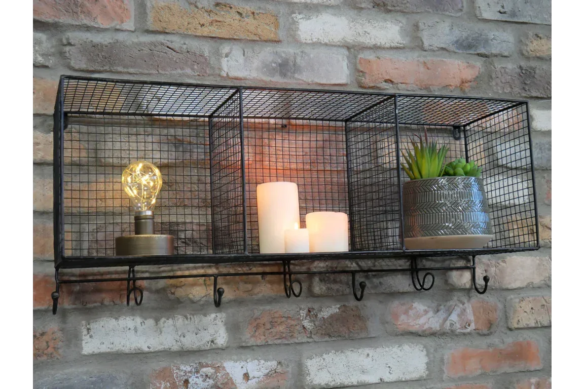 Black wire wall shelf with hooks, holding lamp, candles, and potted plant on brick wall